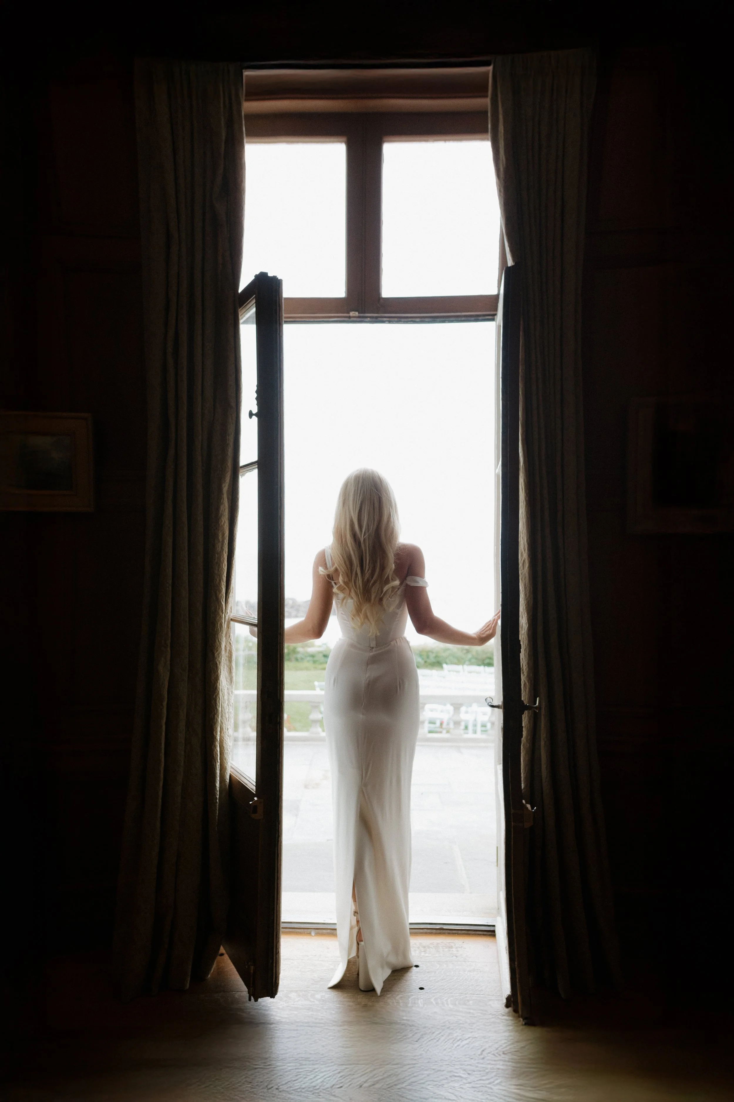 A woman with long blonde hair in a white dress standing at open window, looking outside, with curtains on either side.