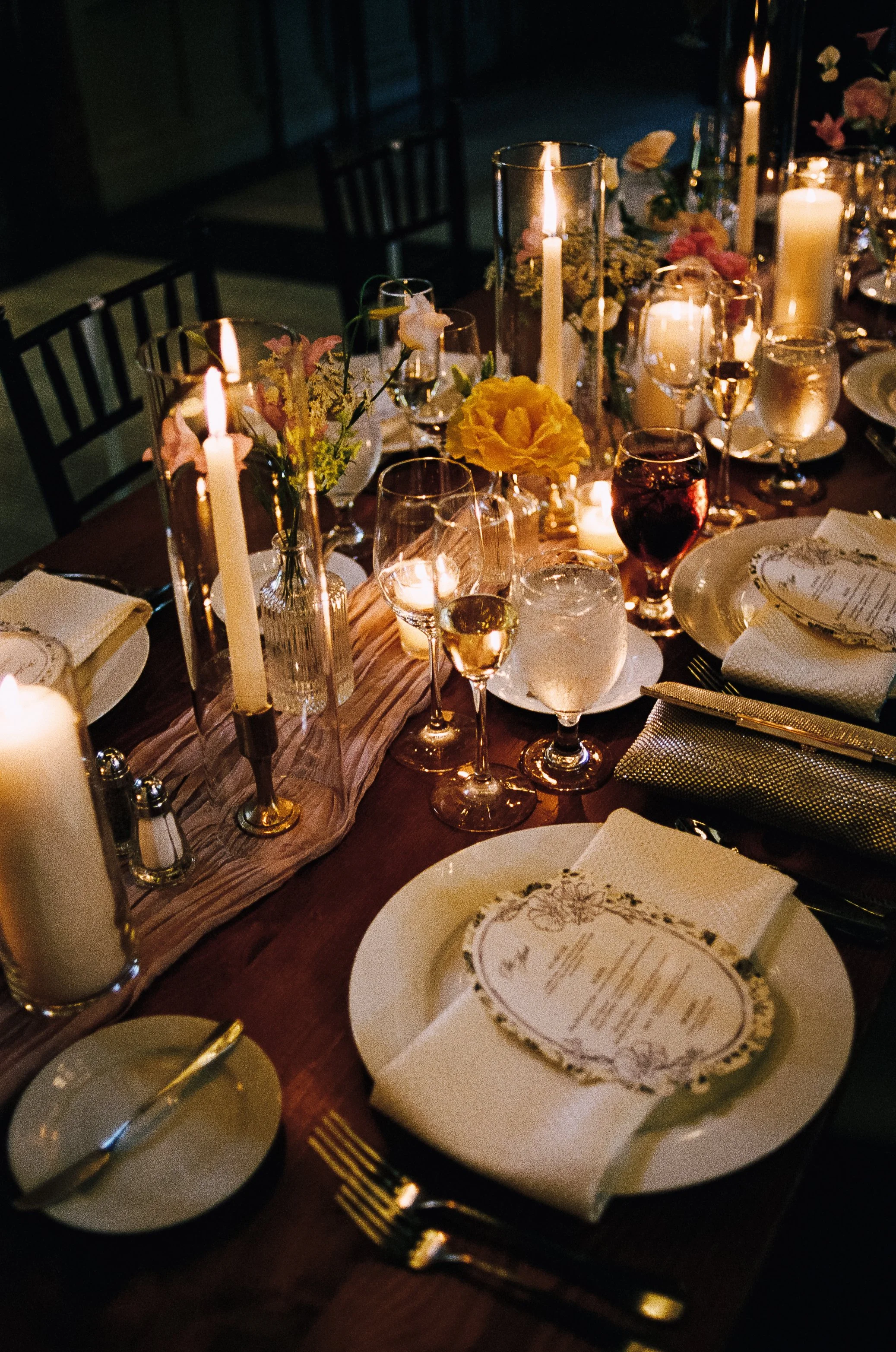An elegant dinner table set with lit candles, floral arrangements, wine glasses, plates with menus, and gold cutlery, creating a sophisticated atmosphere.