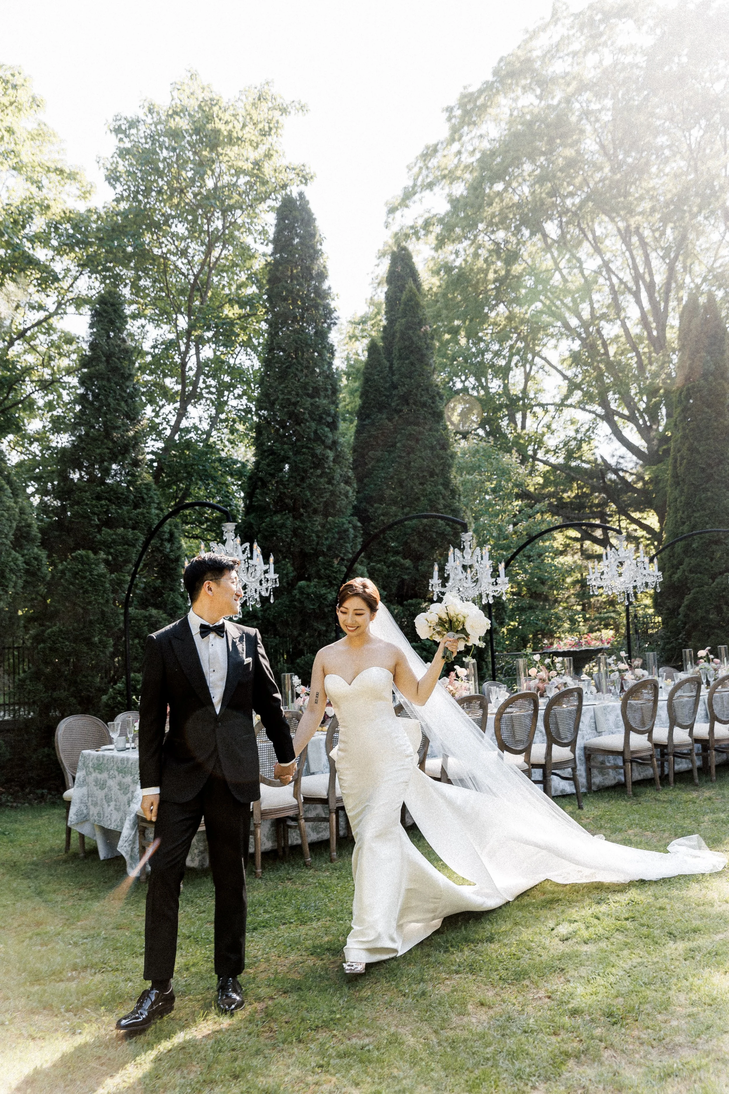 A bride and groom walking hand in hand outdoors during their wedding celebration. The bride is wearing a white strapless wedding gown with a long train and veil, holding a bouquet of white flowers. The groom is dressed in a black tuxedo with a bow ti