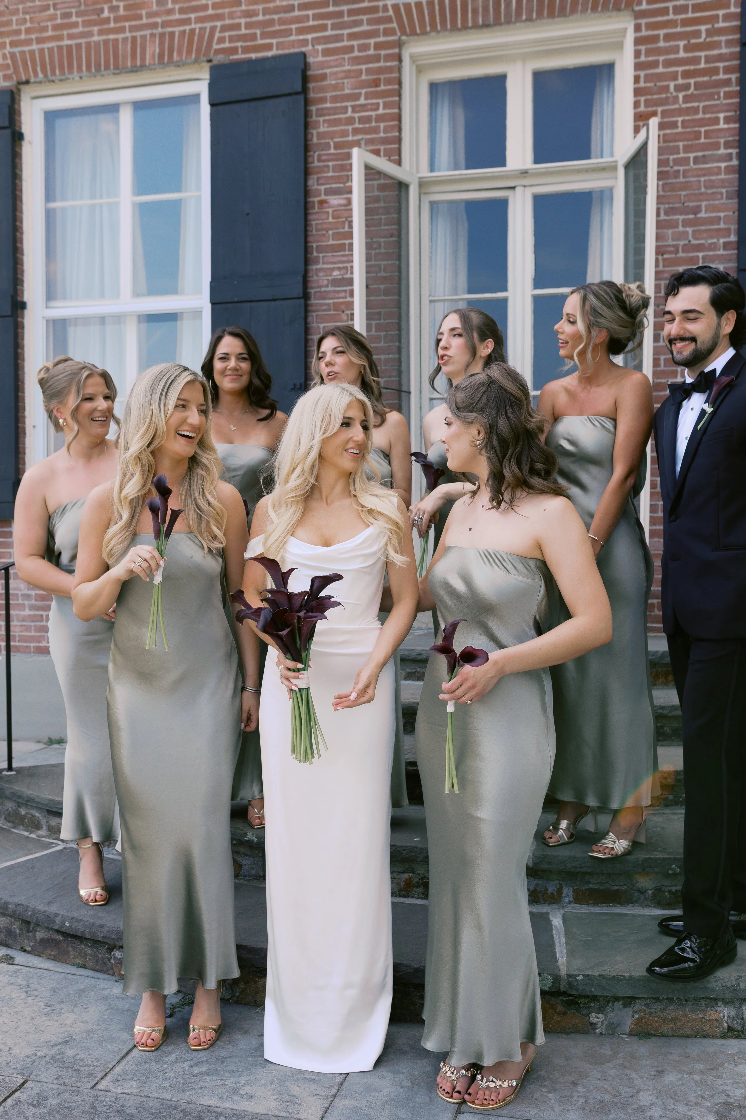Group of women in dresses and one man in suit standing outside, holding dark purple calla lilies, smiling and talking.