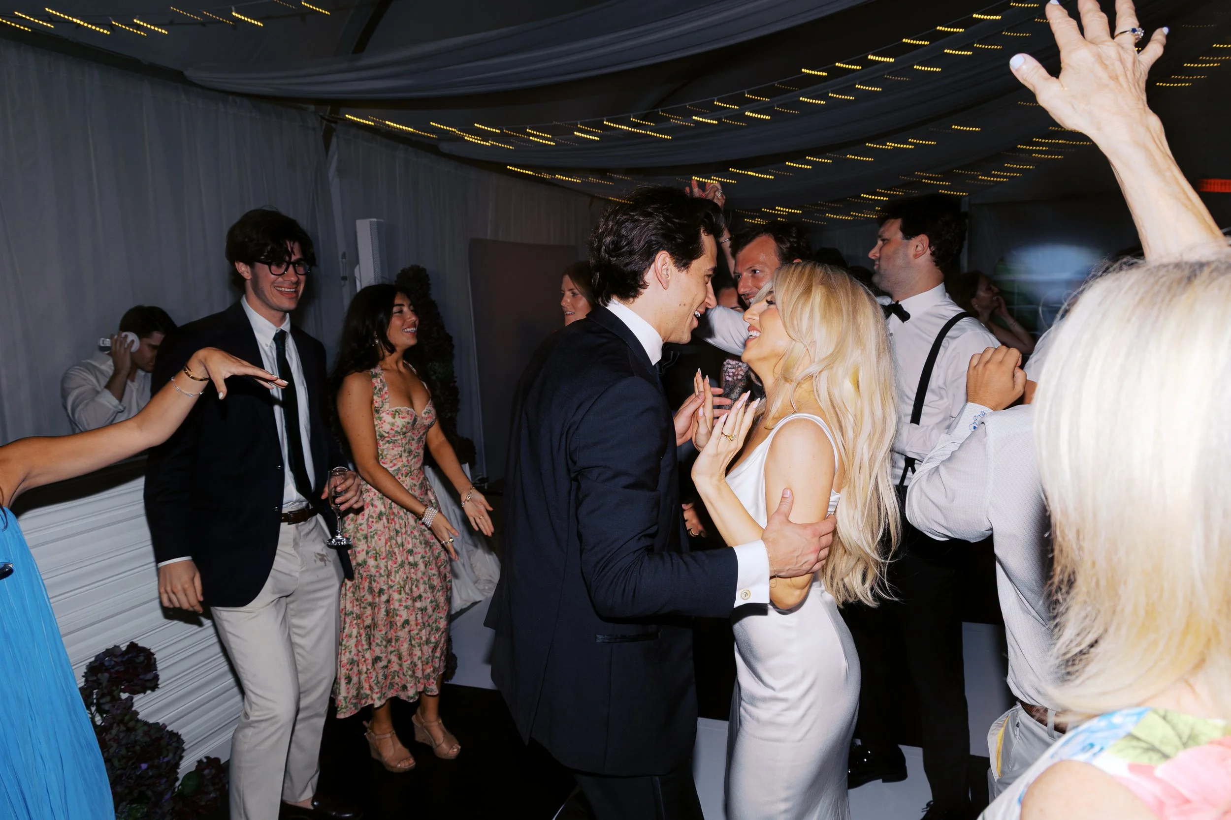 A couple dancing closely at a wedding reception, smiling and gazing at each other, surrounded by friends dancing and celebrating under string lights.