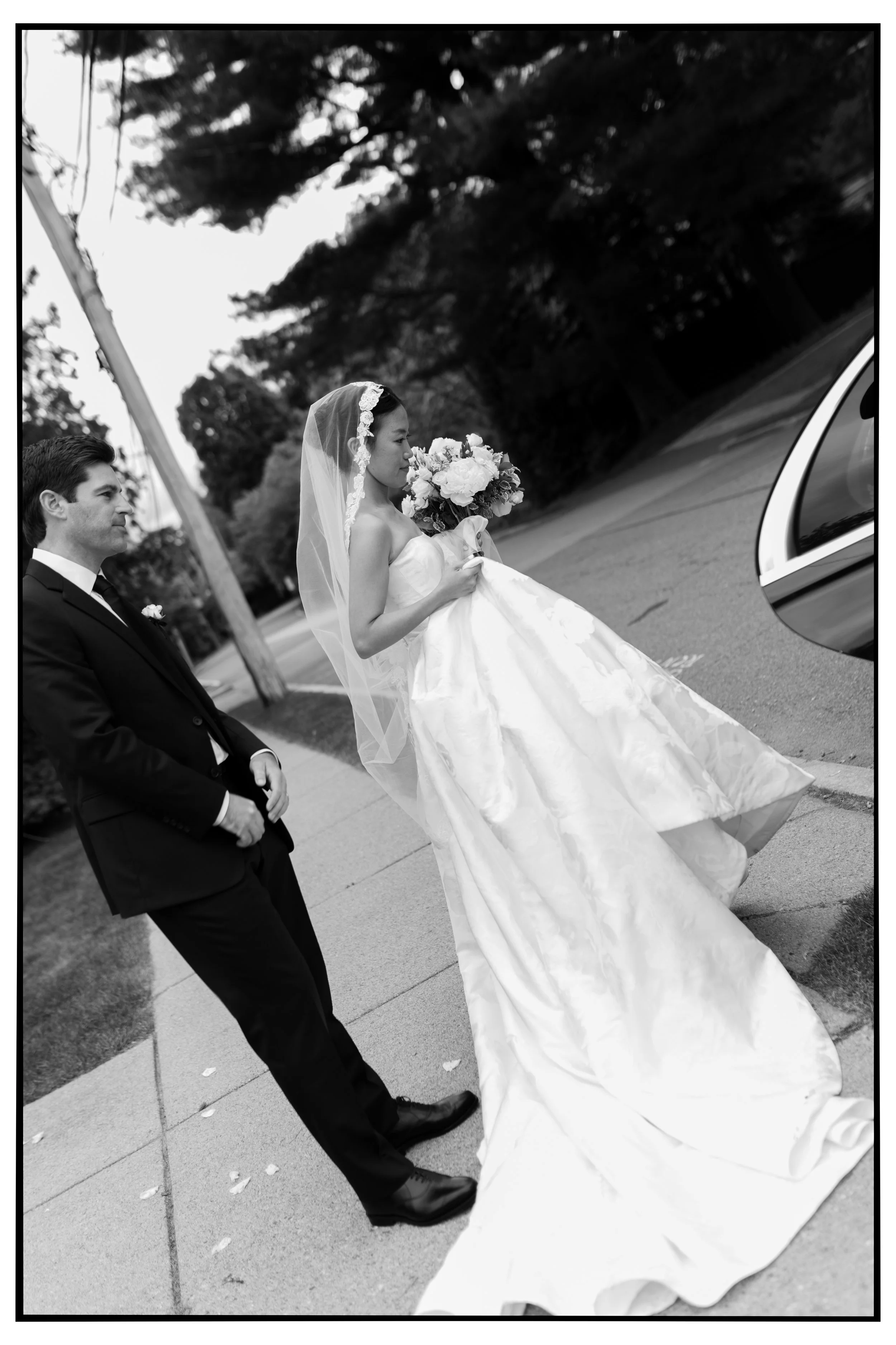A bride in a wedding dress holding a bouquet and a groom in a suit walking on a sidewalk near a parked car, with trees in the background.
