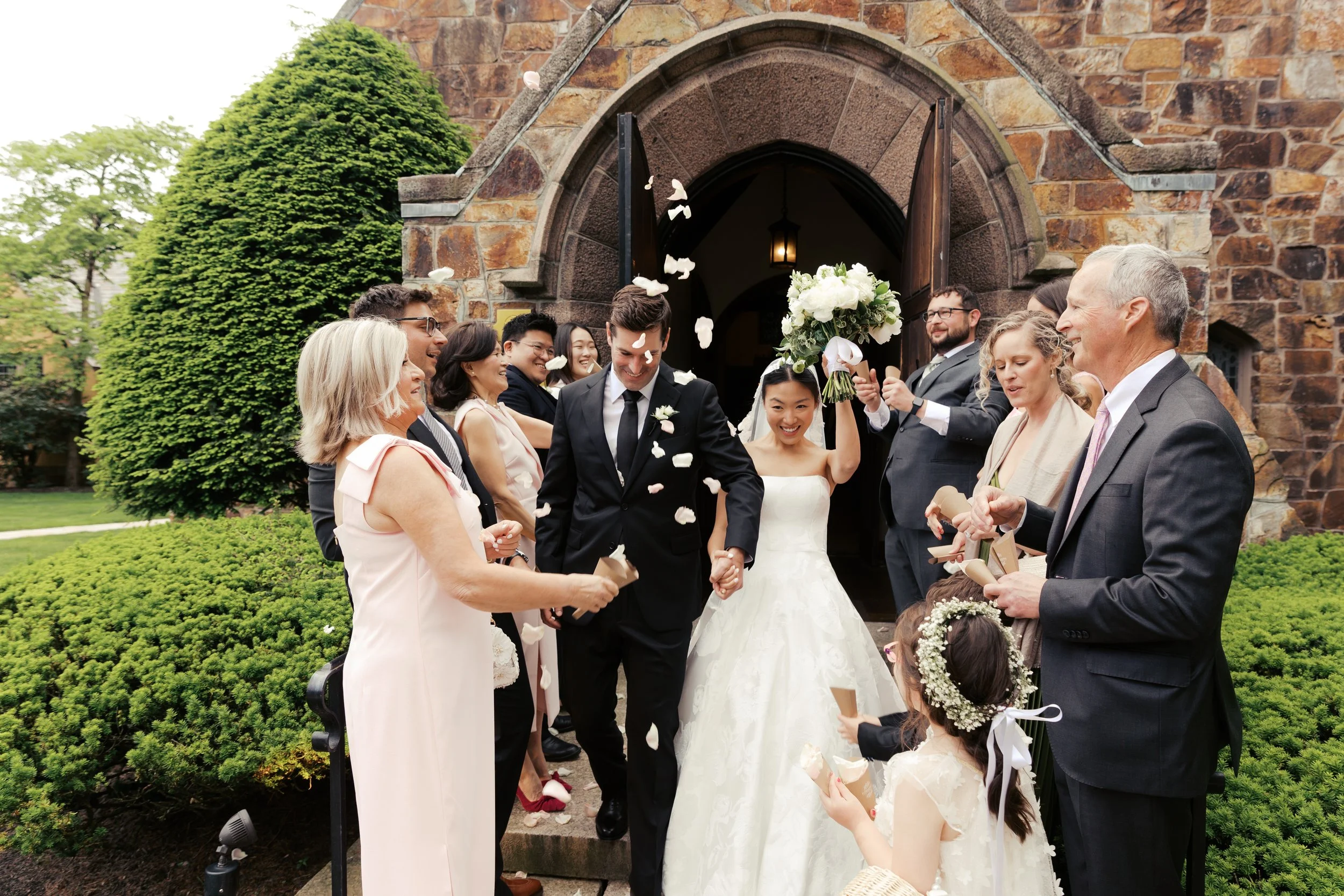 A wedding celebration outside a church with people throwing flower petals as the bride and groom hold hands and smile.