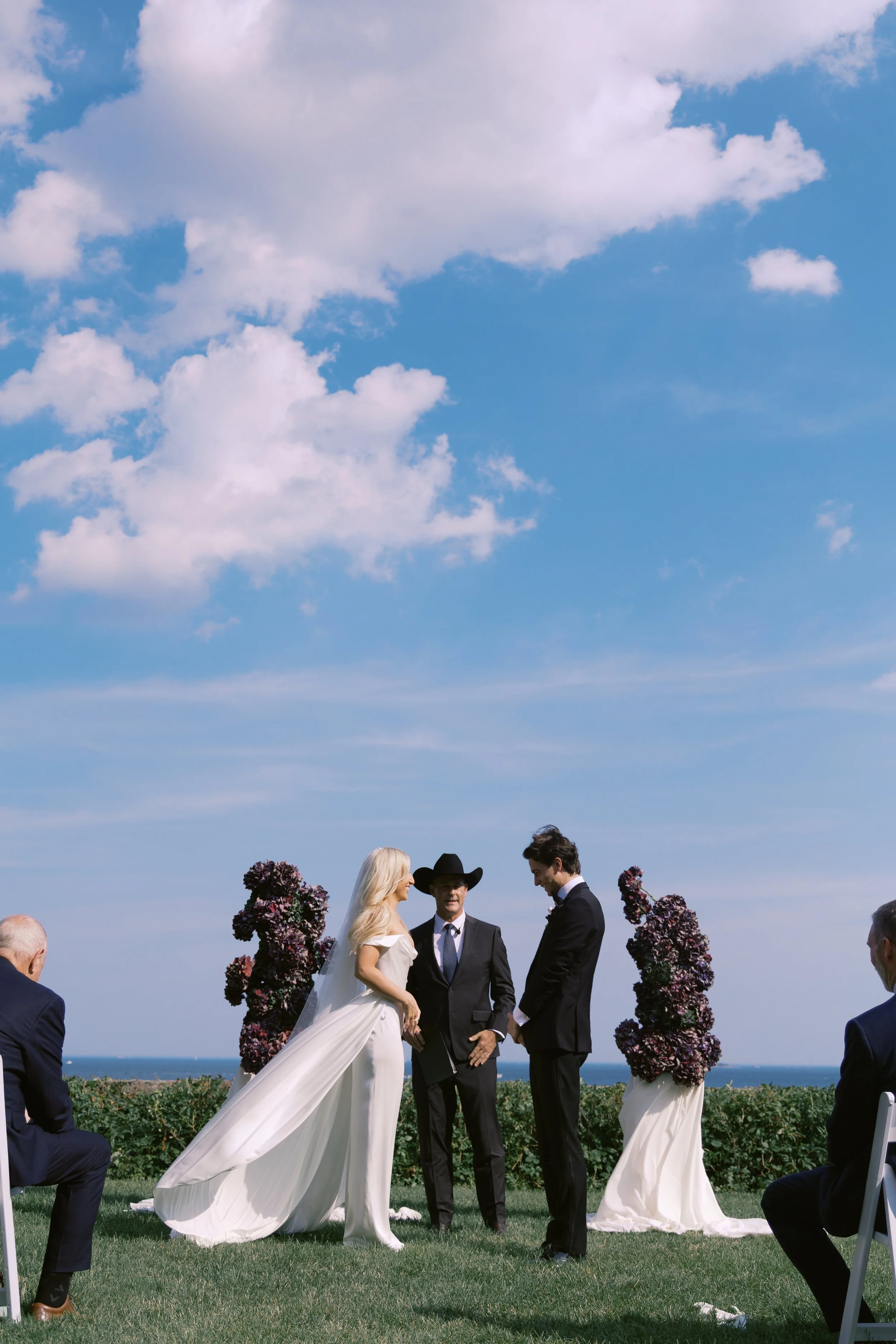 A wedding ceremony taking place outdoors on a grassy area with a view of the ocean and blue sky with clouds. The bride and groom stand facing each other, with an officiant between them, under floral arrangements, while guests sit on either side.