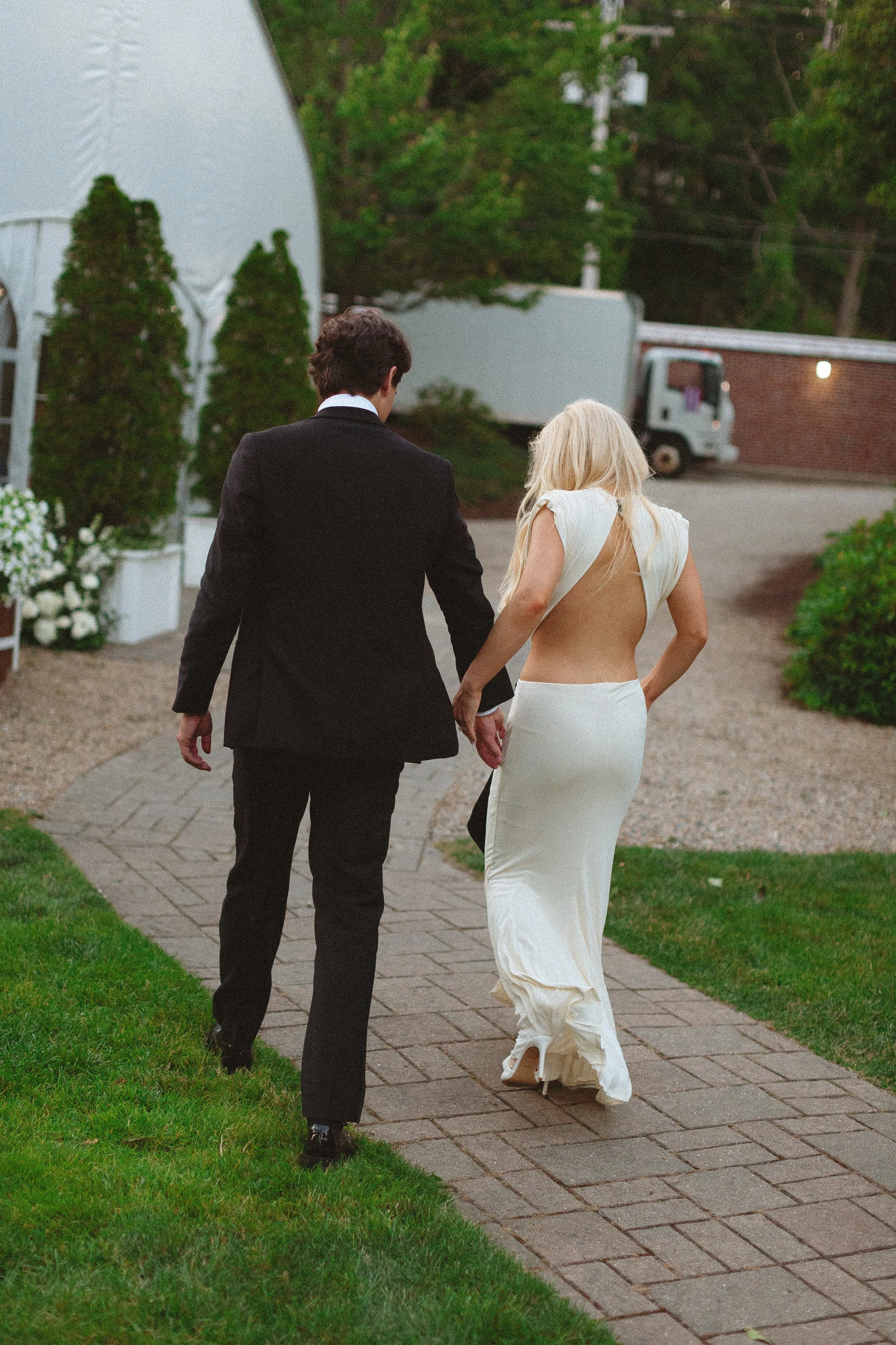 A couple, a man in a black suit and a woman in a white wedding dress, walking hand in hand on a brick pathway outdoors during evening.