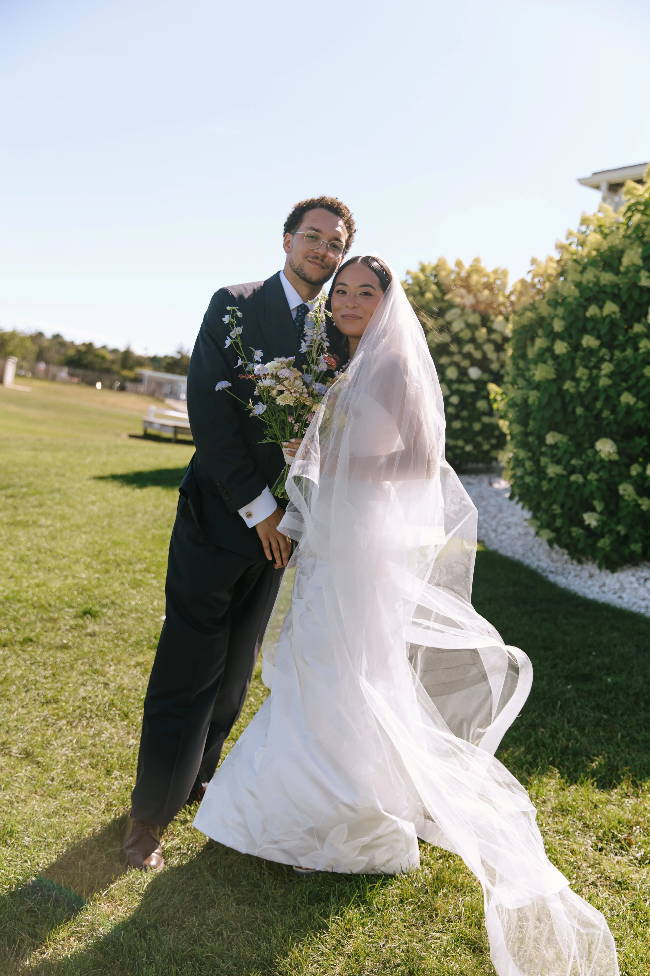 A newlywed couple standing on a grassy lawn, with the bride in a white gown and veil, holding a bouquet, and the groom in a dark suit, smiling at the camera.