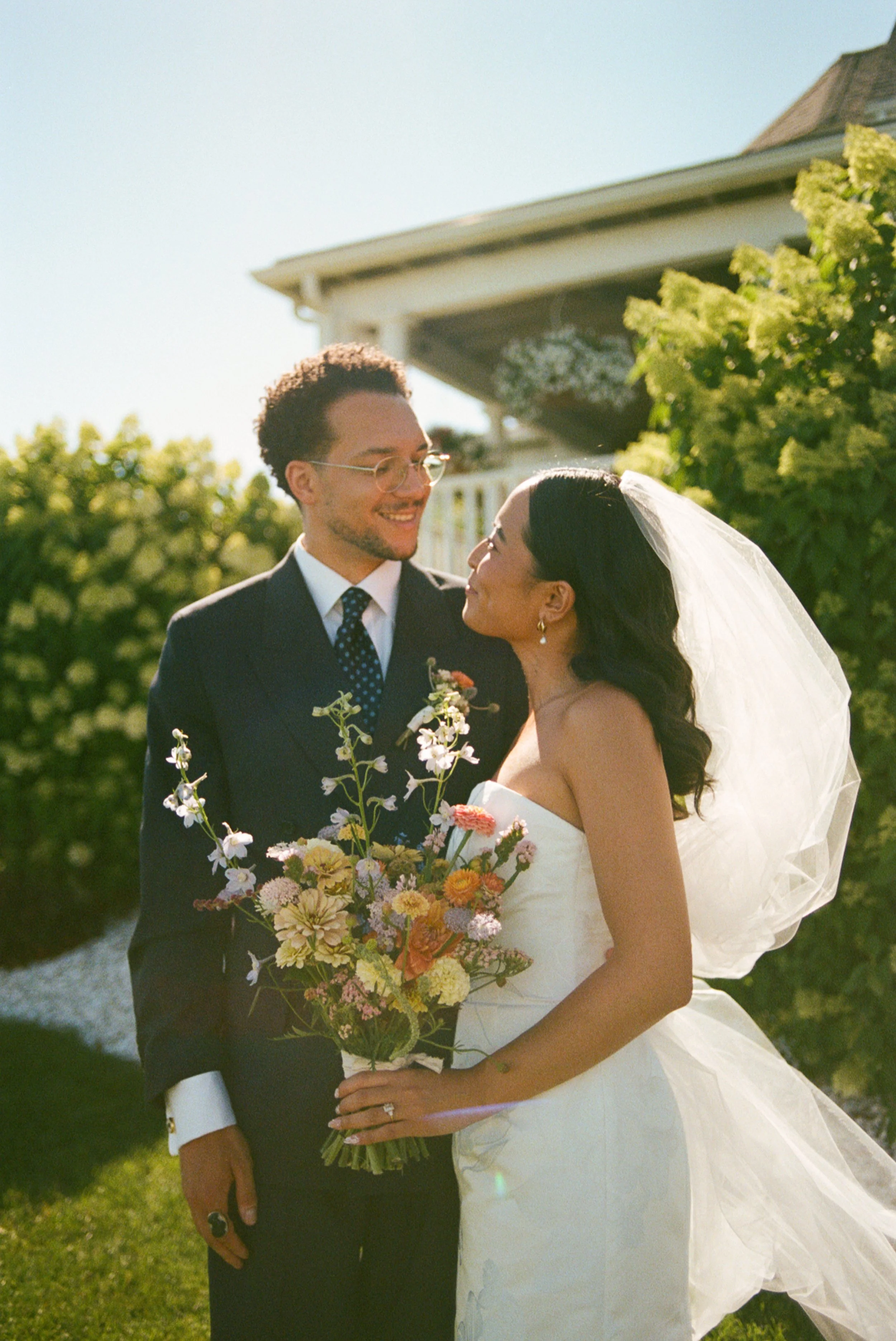 A bride and groom on their wedding day outdoors, smiling and gazing at each other. The bride is holding a colorful bouquet, wearing a strapless white wedding dress and veil. The groom is in a dark suit with a tie and glasses. Background includes gree