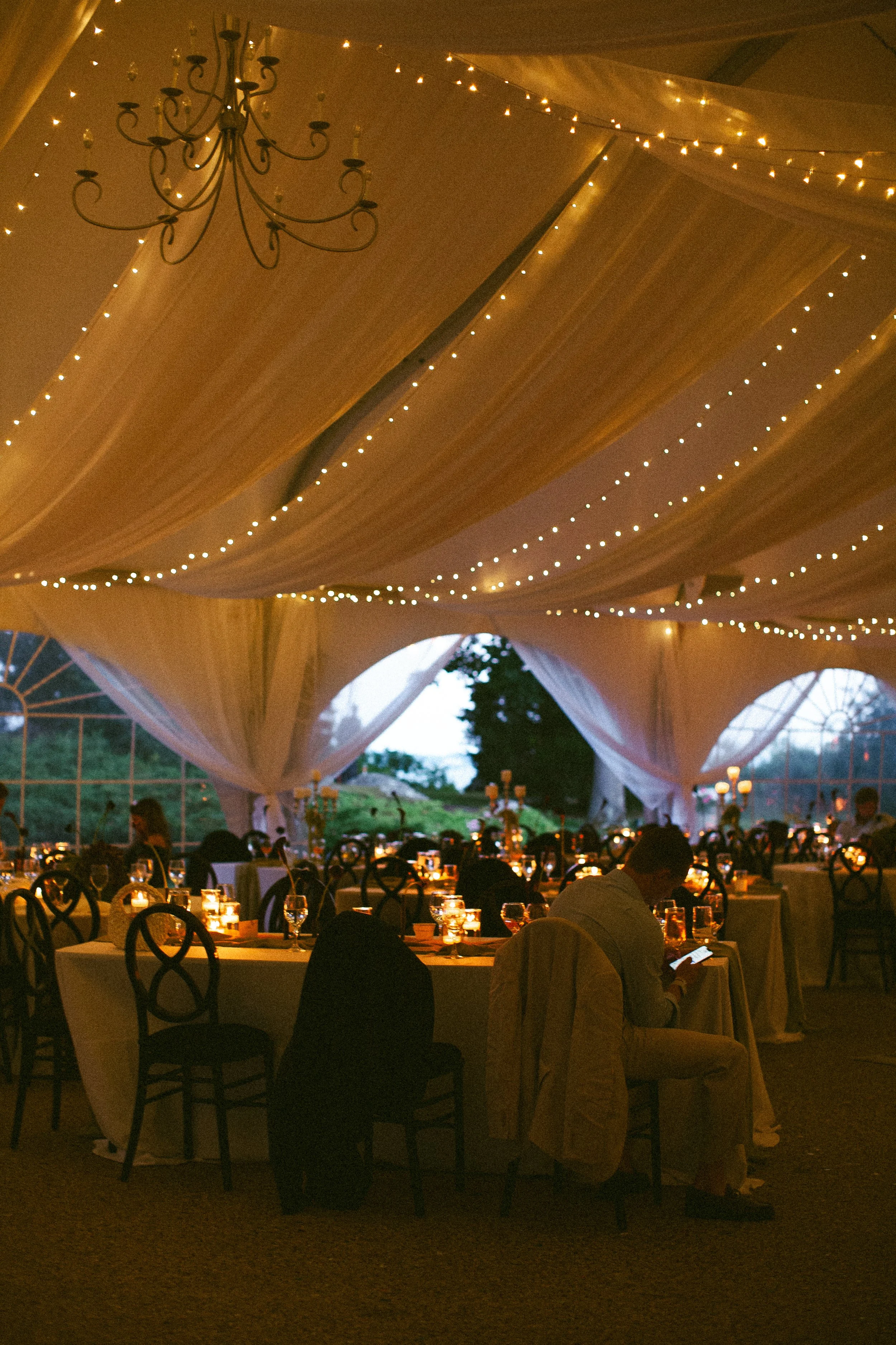 Indoor evening wedding reception under a large tent decorated with string lights and draped fabric, with round tables set with candles and glassware, and guests seated and mingling.