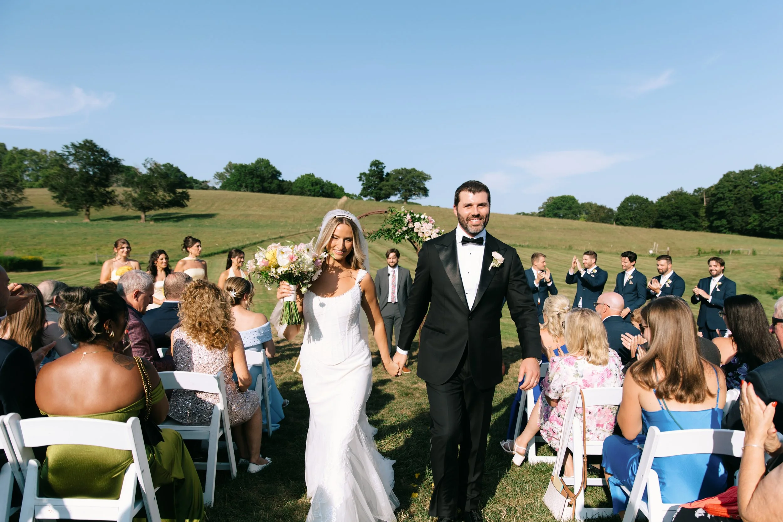 Barn at Gibbet Hill Groton, A bride in a white wedding dress and a groom in a black tuxedo holding hands, walking down the aisle at an outdoor wedding ceremony on a sunny day with guests seated and congratulating them.