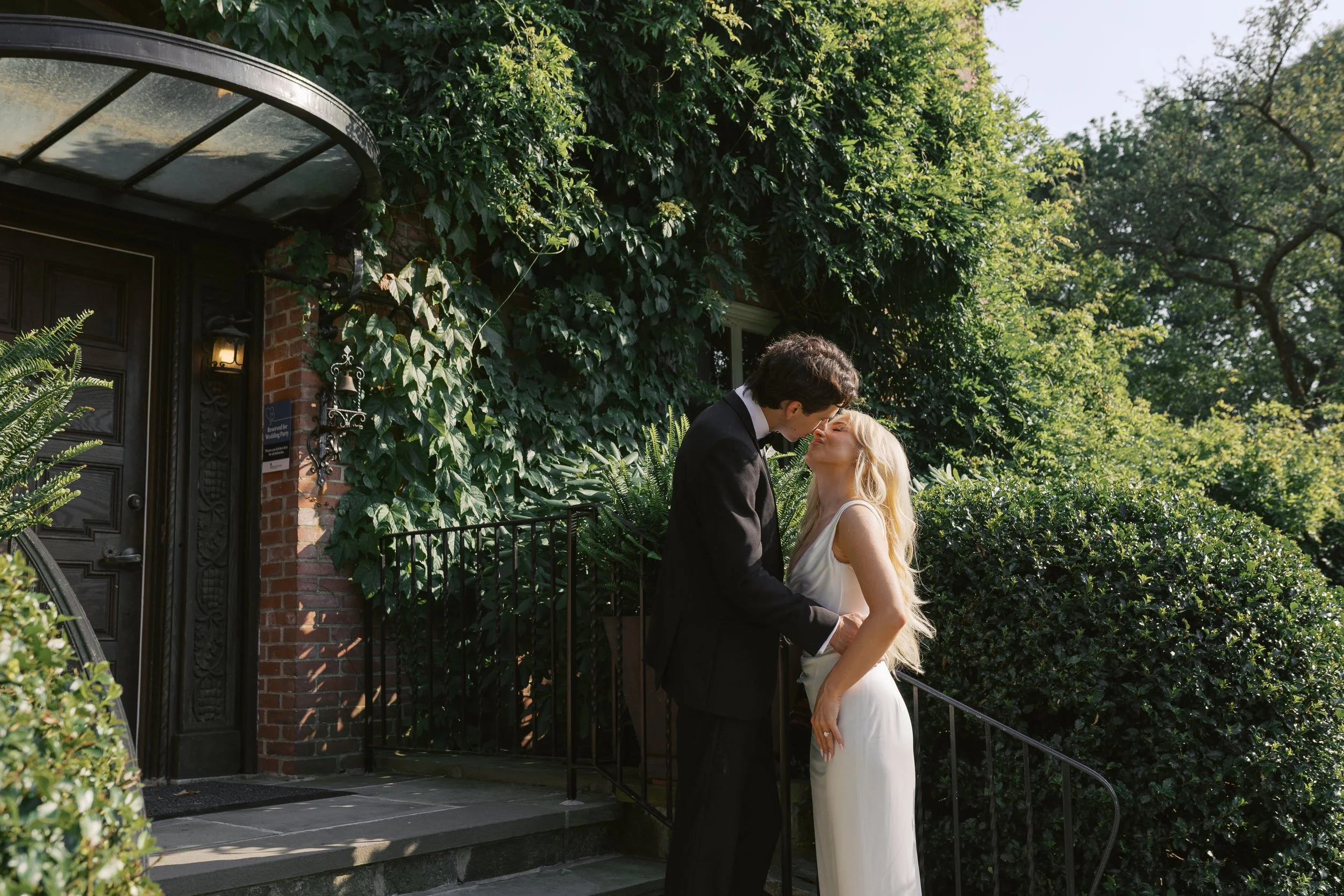 A couple in wedding attire sharing a kiss outdoors in front of a house covered in ivy and greenery.