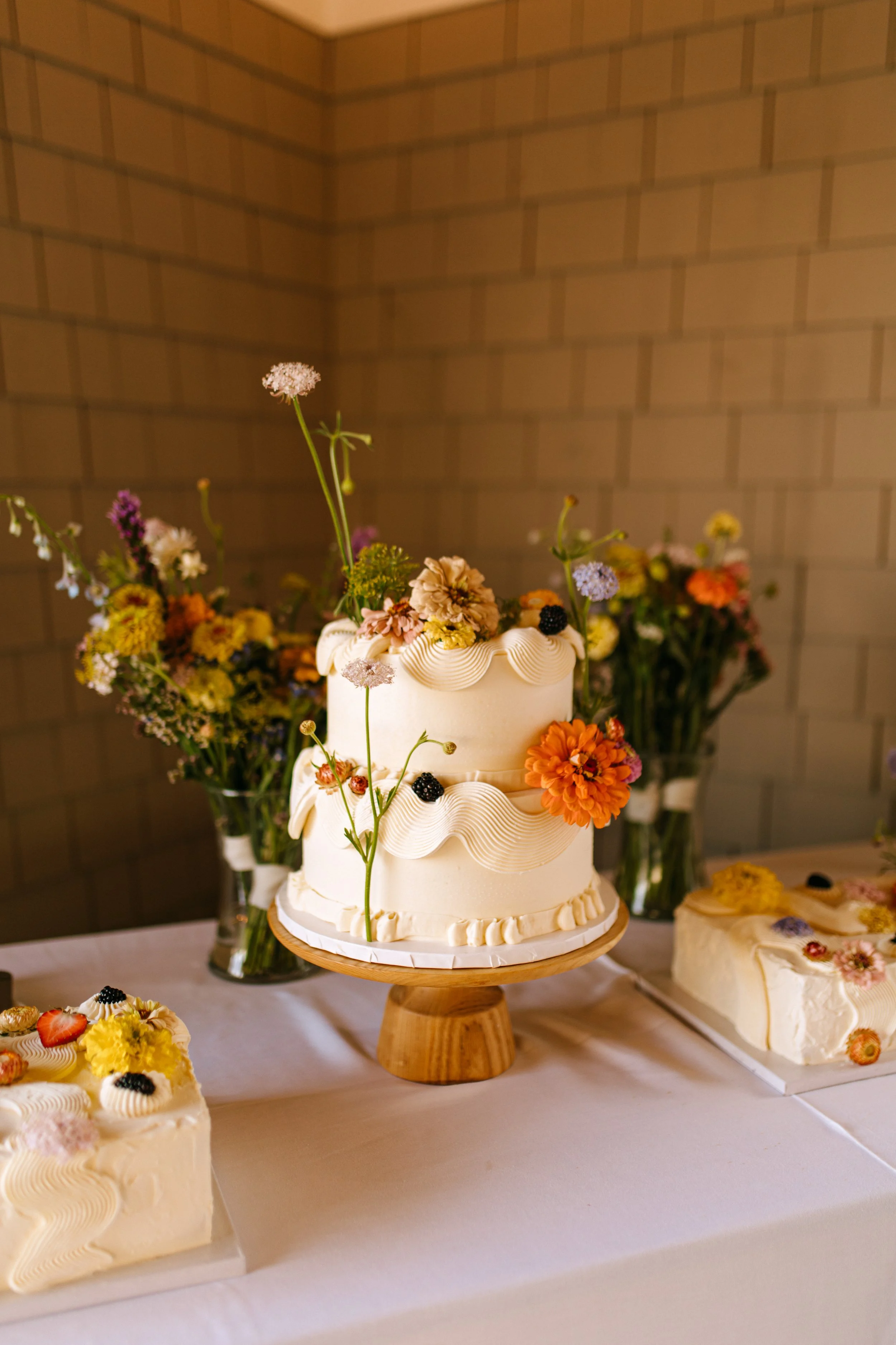 A white, two-tiered wedding cake decorated with fresh flowers and blackberries, placed on a wooden cake stand, with two additional cakes and colorful flower arrangements in the background.