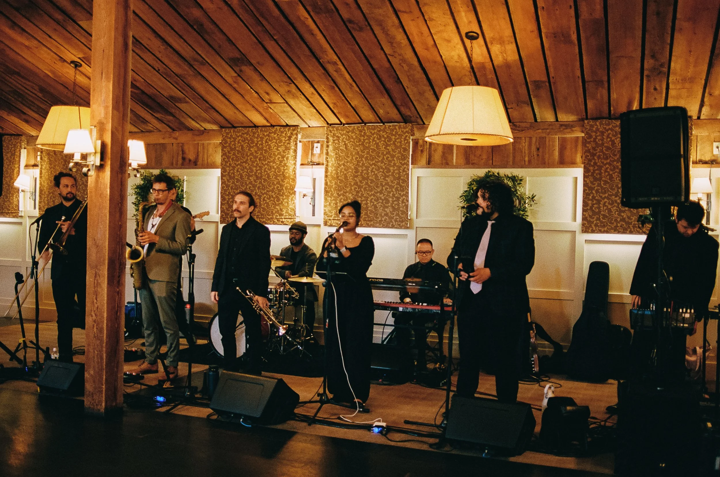 Band performing live in a cozy, warmly lit venue with wooden walls and ceiling, featuring seven musicians with various instruments, including saxophones, keyboard, drums, and microphones.