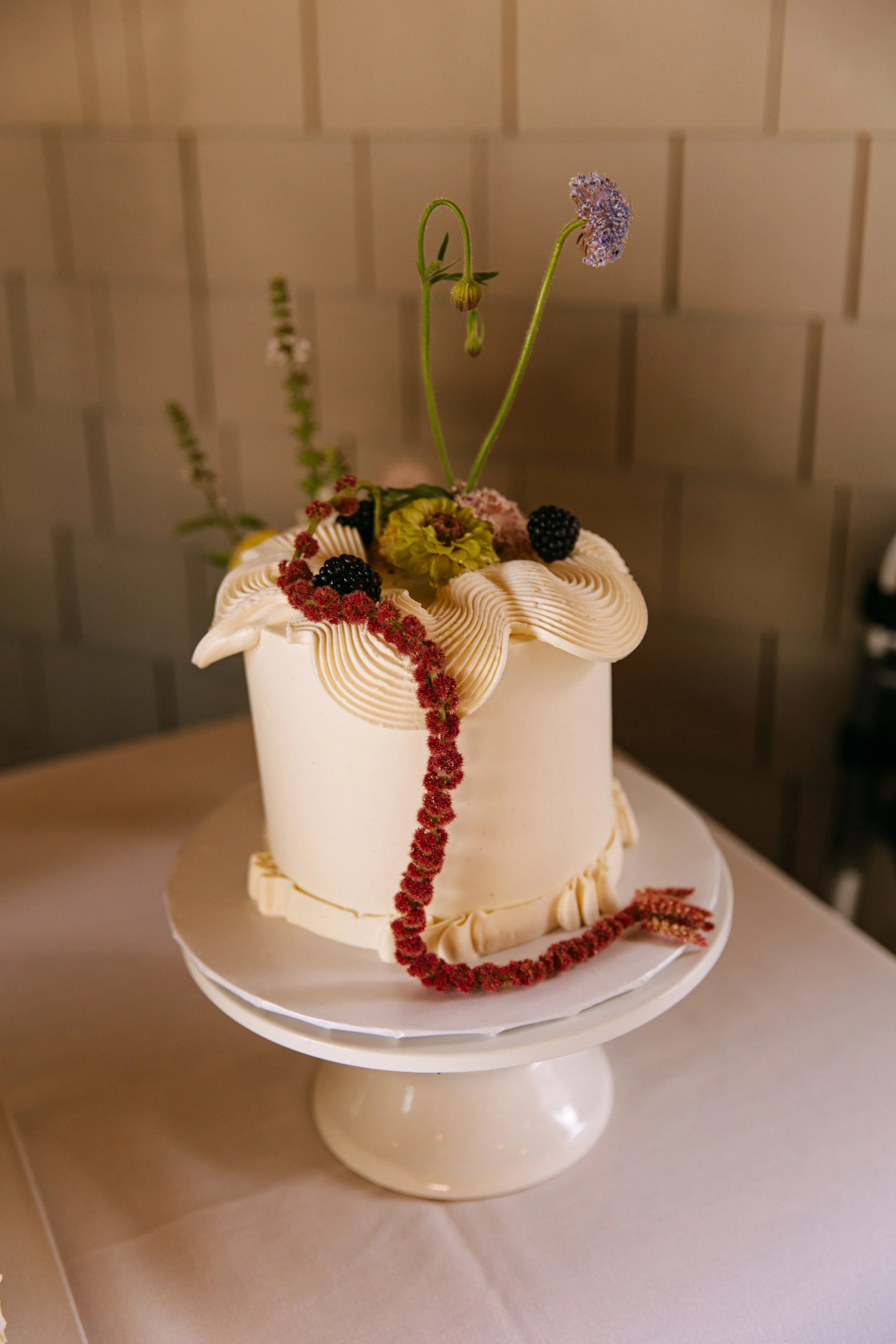 A white cake decorated with cream ridges, blackberries, and red berries, topped with flowers and long green stems, on a white cake stand.