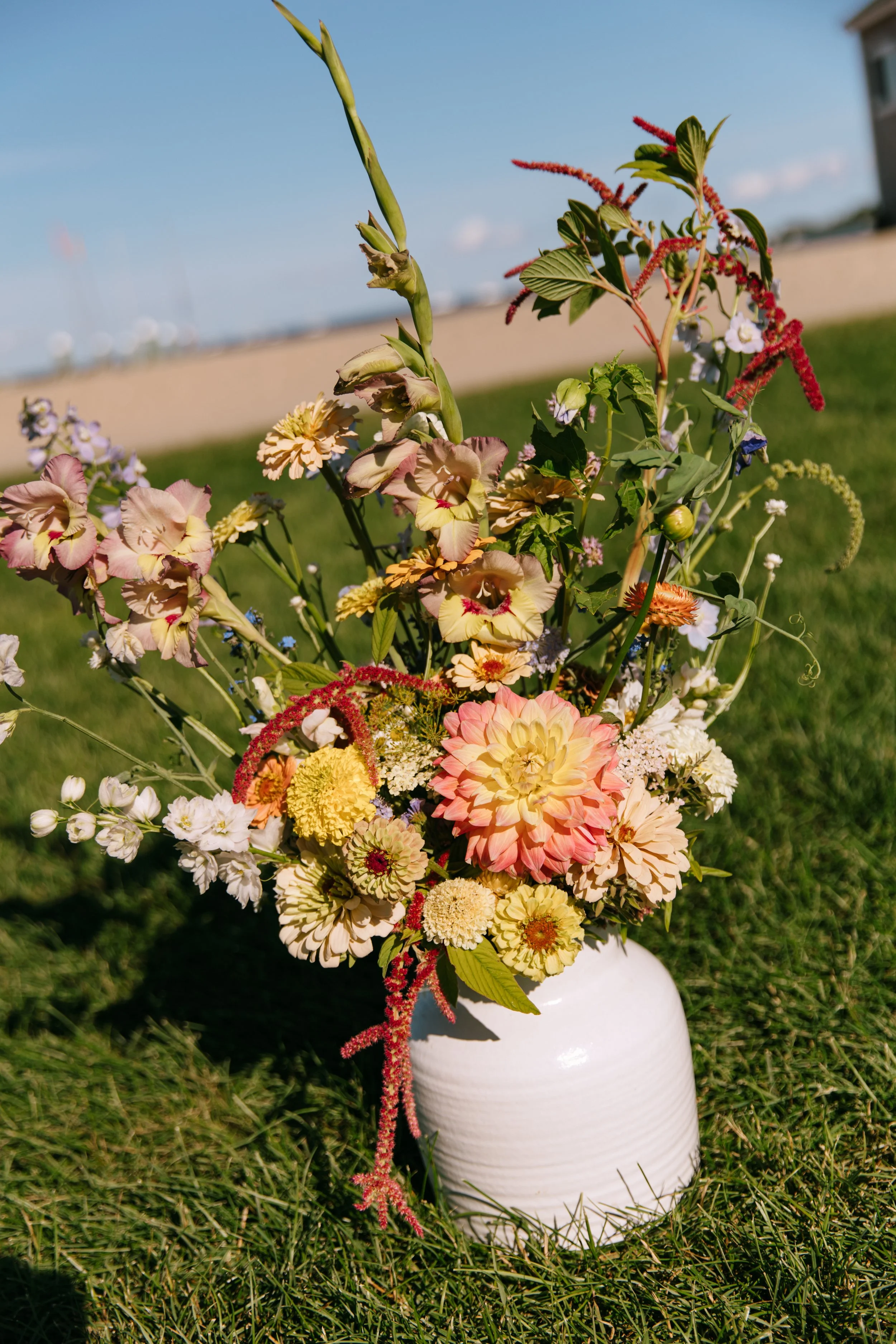 Colorful bouquet of flowers in a white vase outdoors on green grass with a blue sky and blurred background.