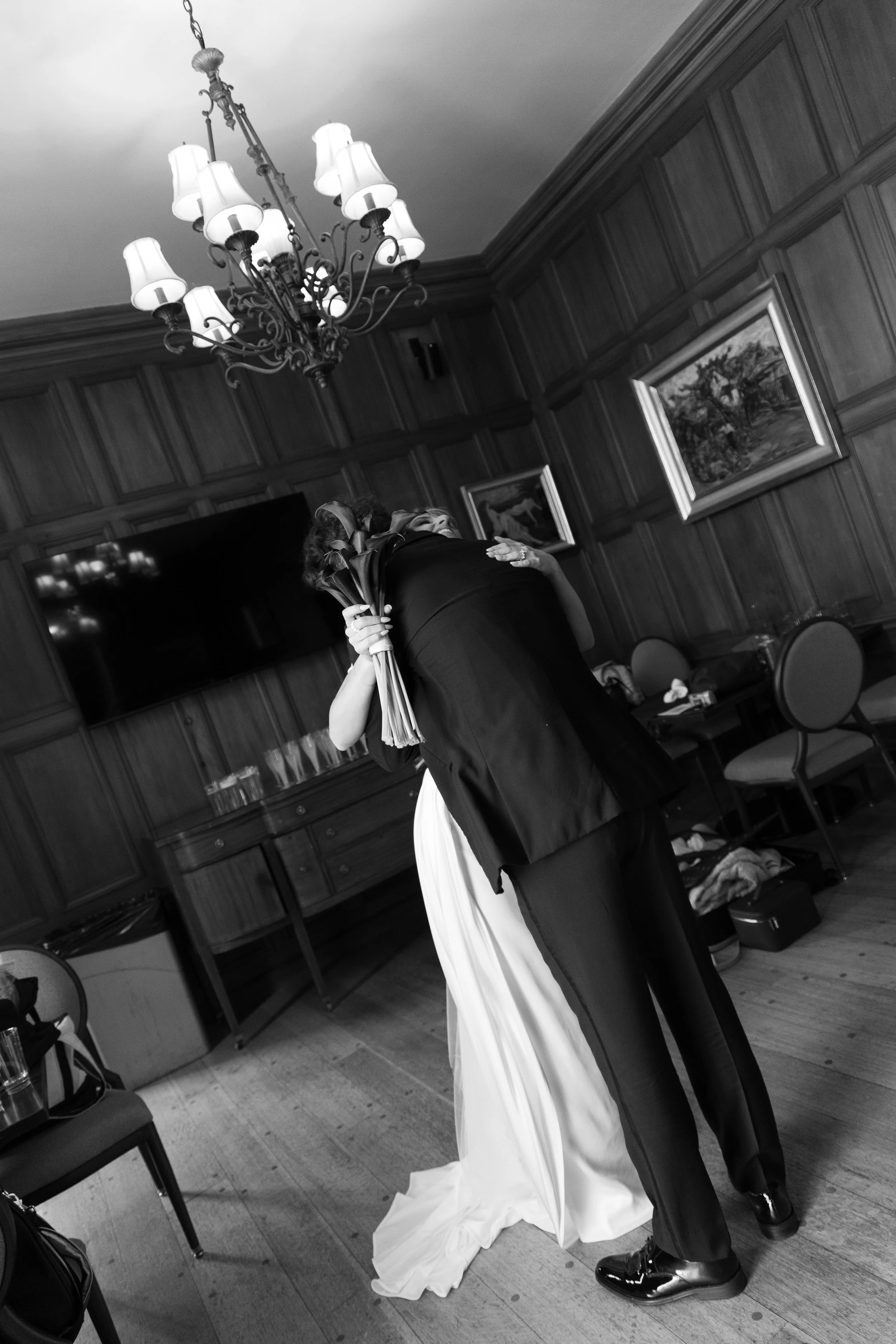 A bride and groom sharing a dance in a wood-paneled room, with a chandelier overhead and framed paintings on the wall.