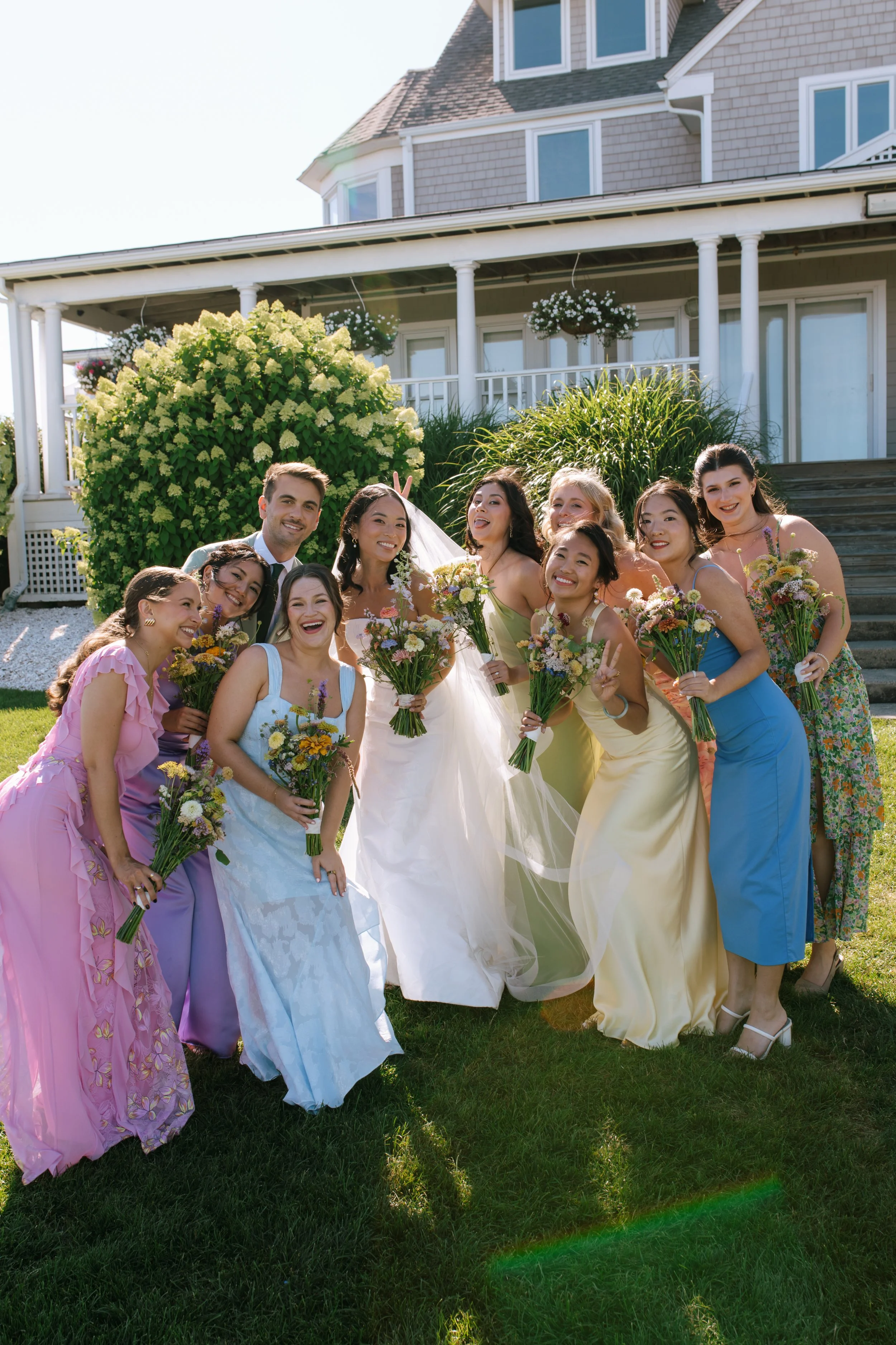 Group of women and a man in wedding attire posing outdoors in front of a house, holding bouquets of flowers.