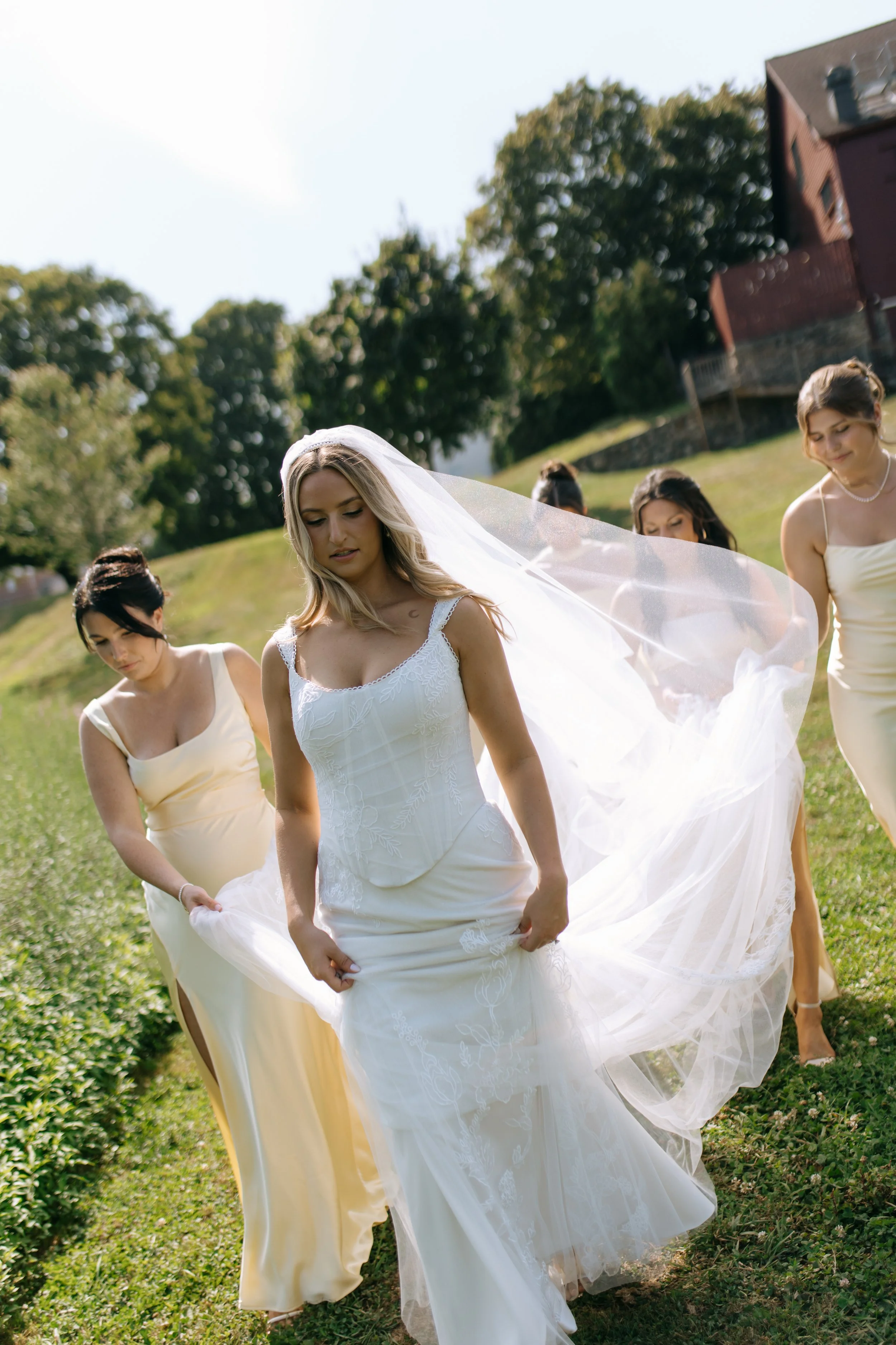 A bride in a white wedding dress and veil walking outdoors on grass, surrounded by bridesmaids in light-colored dresses.