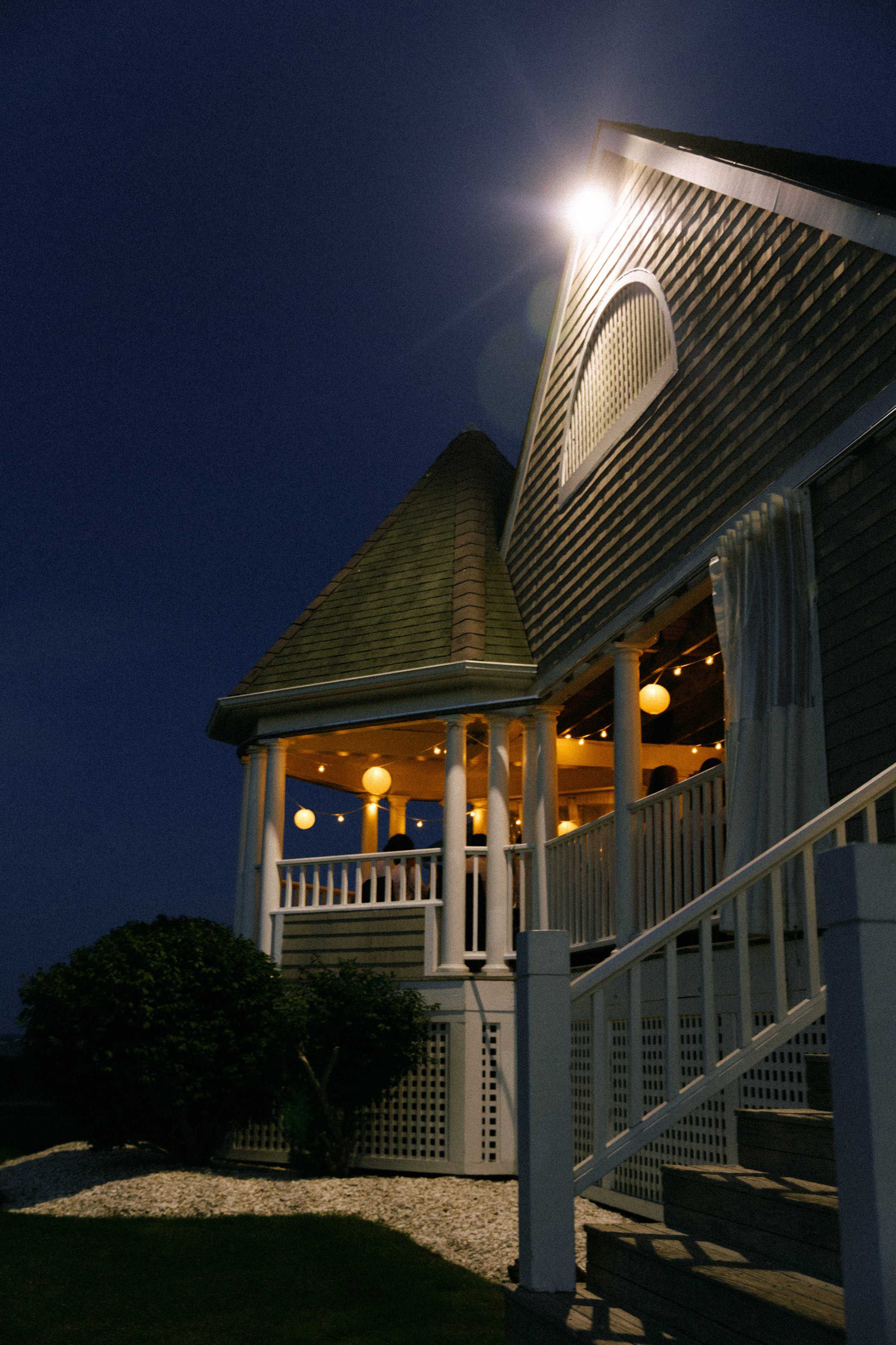 Night view of a house with a turret, a porch with string lights and paper lanterns, and a tree in the foreground.