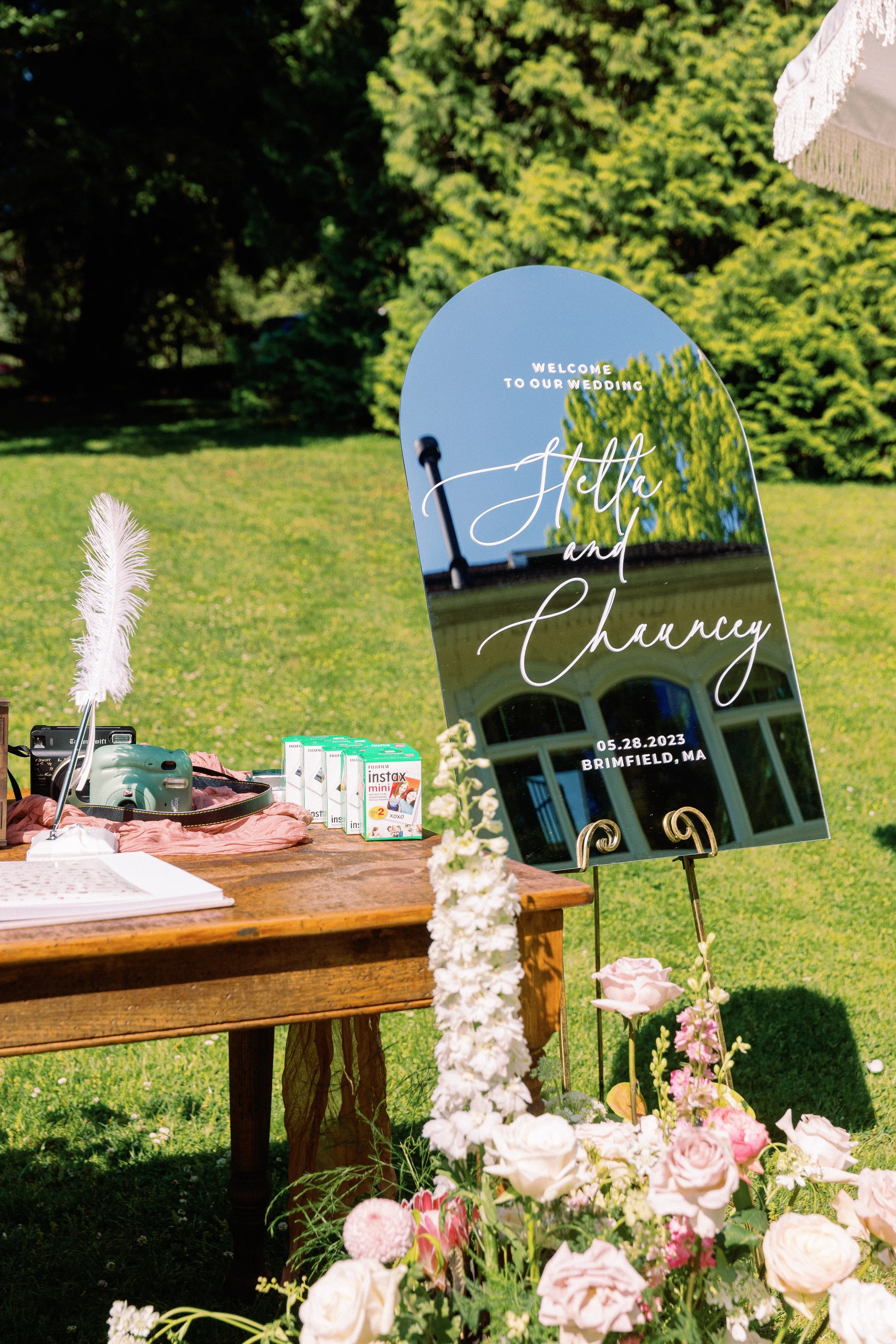 A mirror with handwritten text that reads Welcome to our wedding, Helle and Chancy, 05.28.2023, Brimfield, MA, is displayed outdoors on a grassy lawn, decorated with pink and white flowers, alongside a table with some boxes of instant cameras and a s