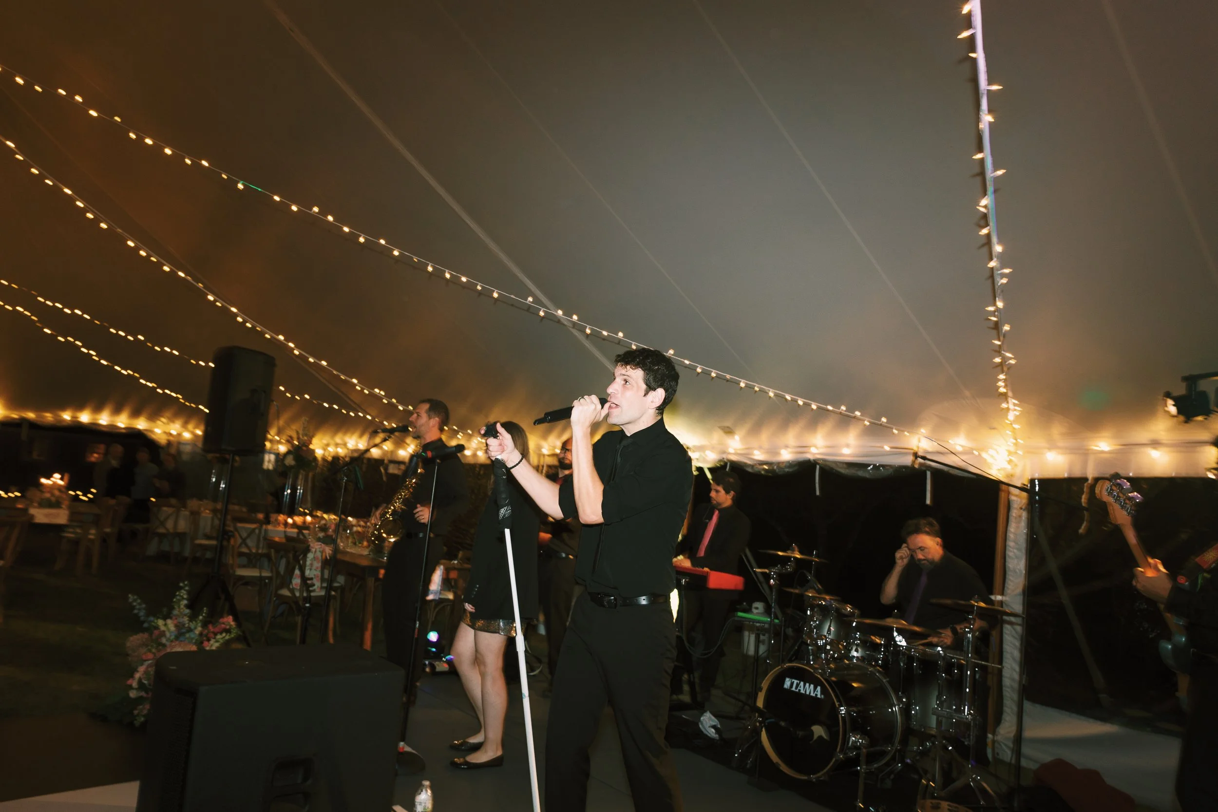 A band performing under a large tent with string lights, featuring a lead singer holding a microphone, musicians with keyboard, drum set, and saxophone, and an audience sitting at tables.