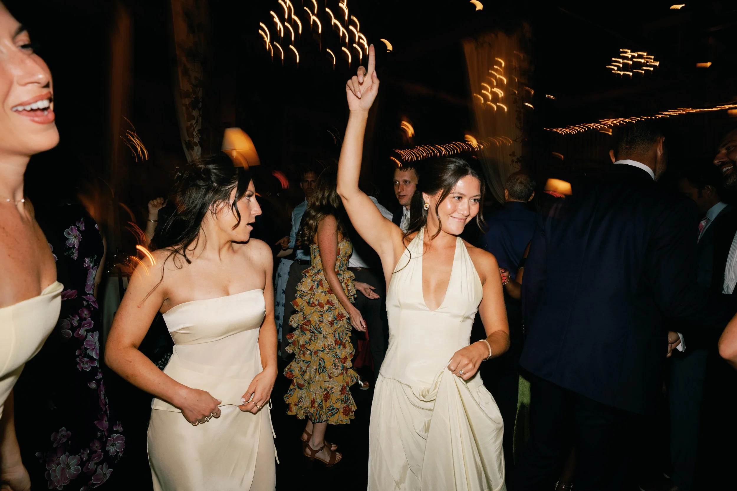 People dancing at a wedding reception, including a woman in a cream dress raising her arm and smiling, with other guests dancing around her in a dimly lit room with string lights.