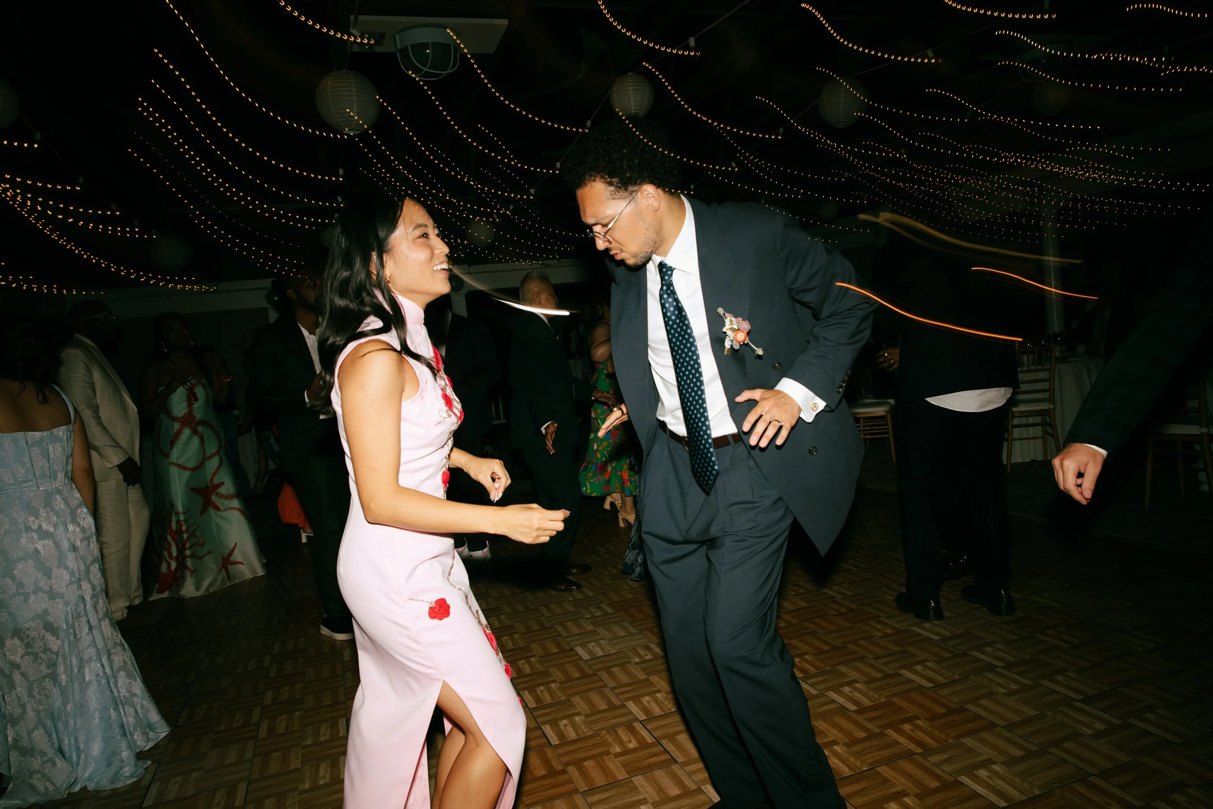 A woman and a man dancing at a celebration or party with string lights overhead, surrounded by other people.