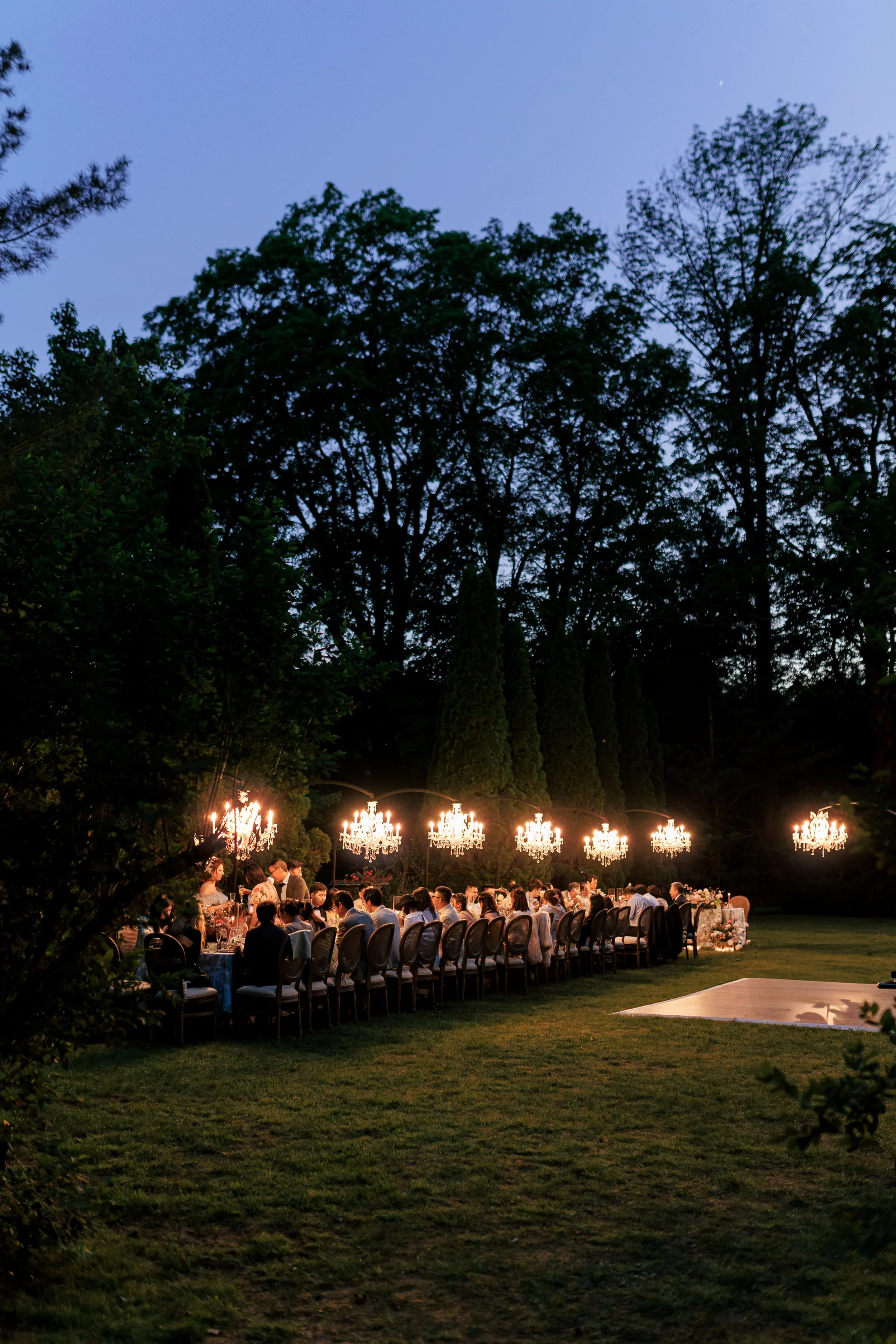 An outdoor evening gathering with a long dining table, chandeliers hanging above, and surrounded by trees and greenery.