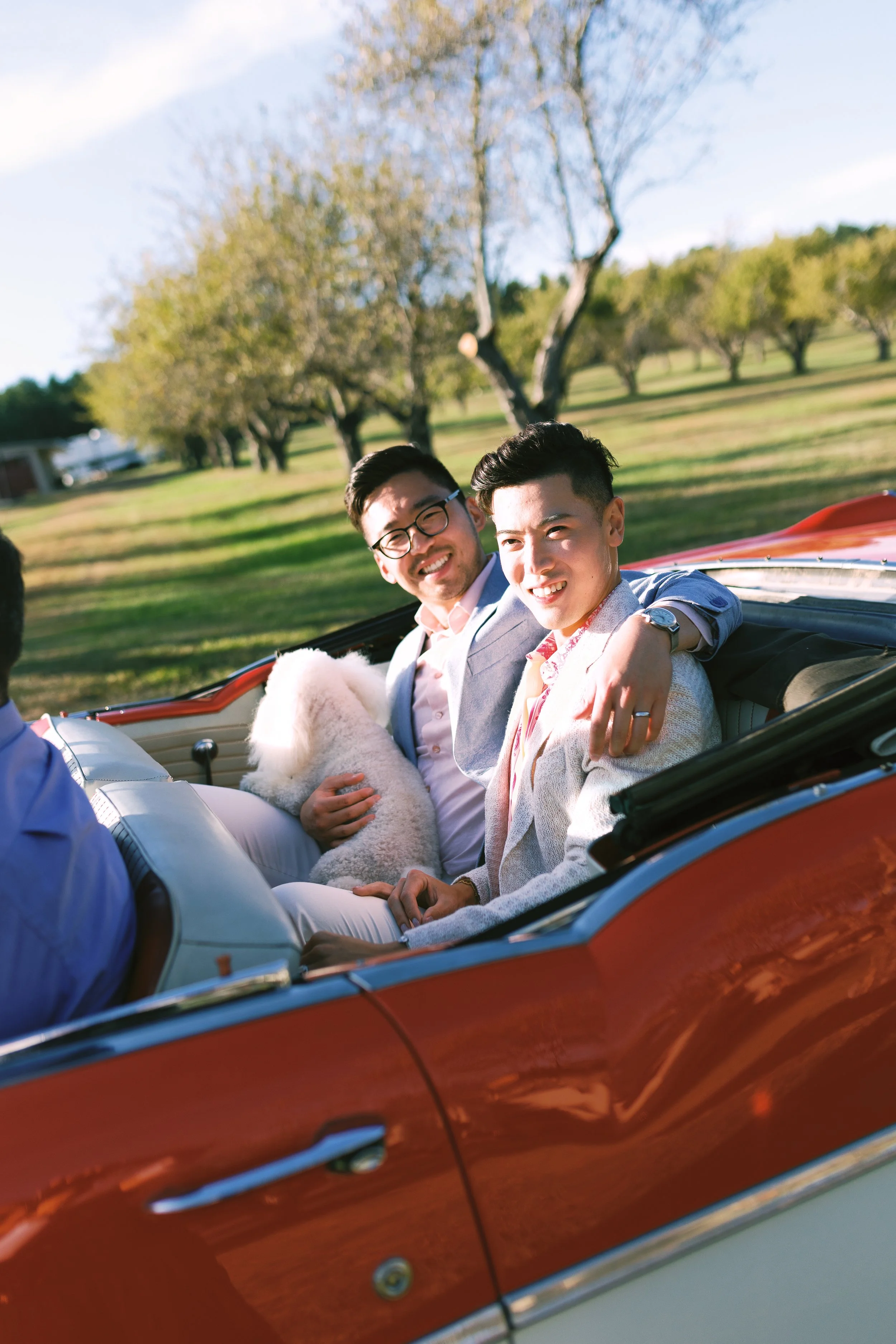 Two young men in formal attire sitting in a red vintage convertible car, smiling, with a fluffy white dog on one man's lap, in a park with trees and clear sky.