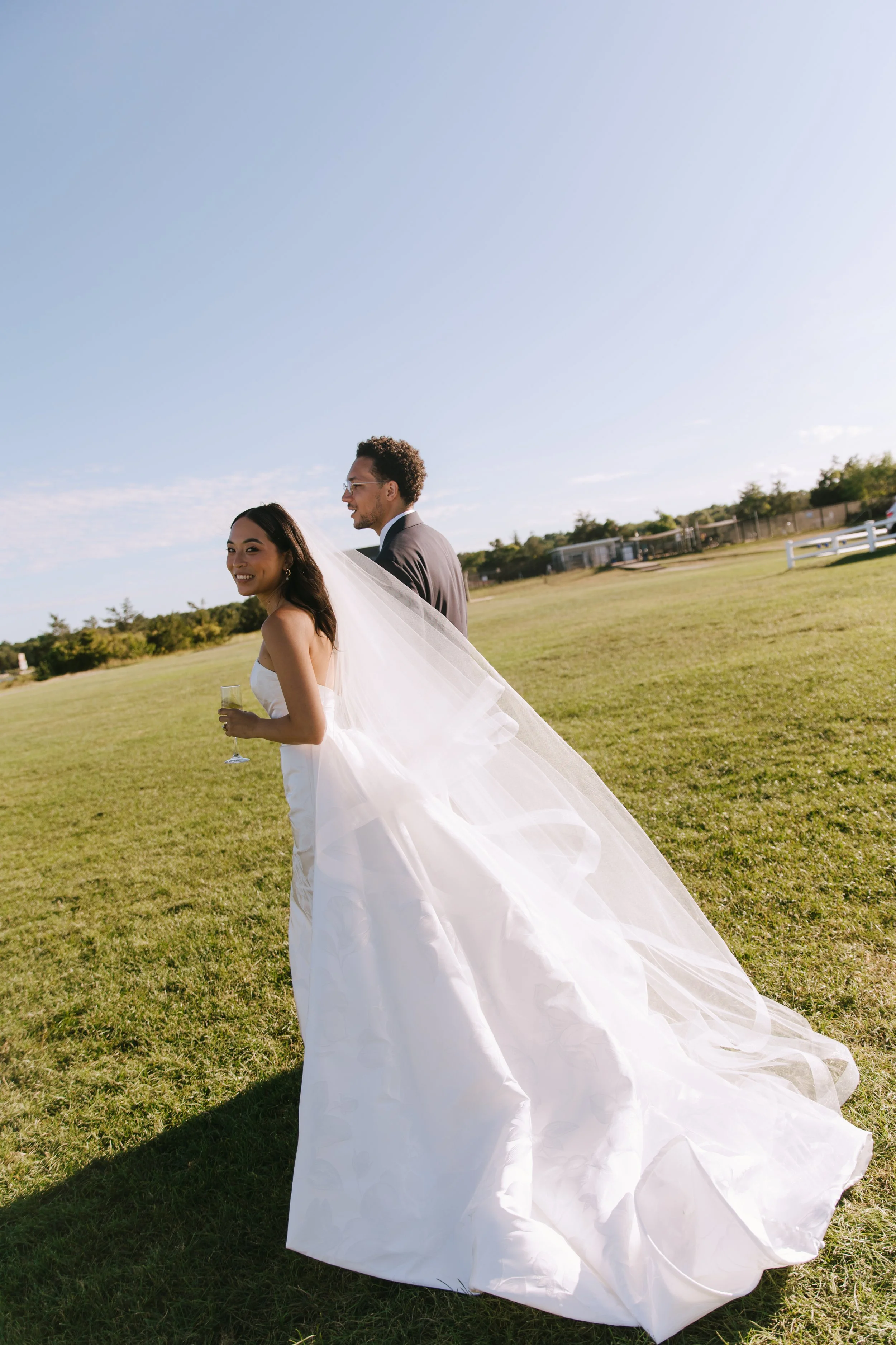 An outdoor wedding scene with a smiling bride and groom standing on grass, with the bride in a white gown and veil holding a glass, and the groom in a suit, under a bright blue sky.