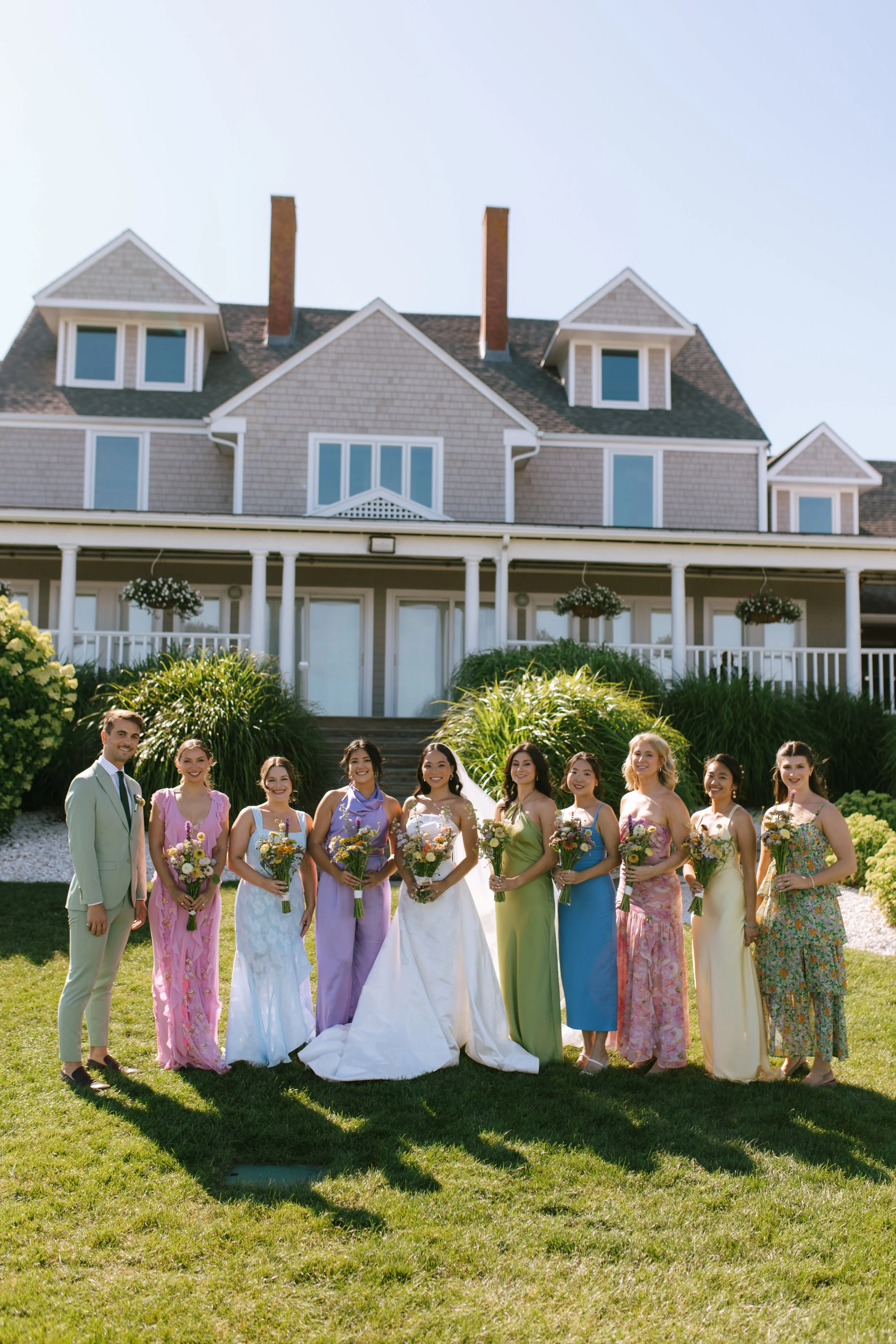 Group of people, mostly women in colorful dresses and one man in a suit, standing outdoors on grass in front of a large house, holding bouquets of flowers, smiling.