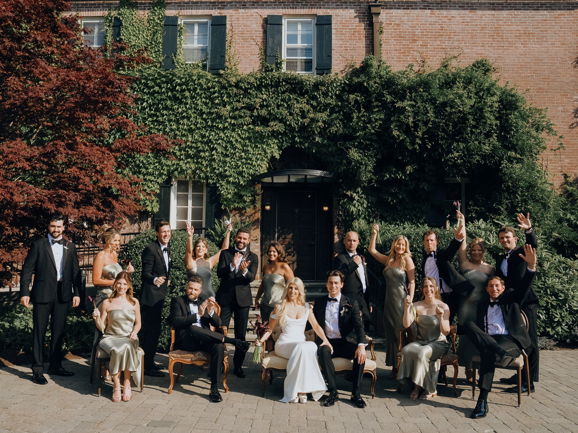 A wedding party of 16 people, including a bride and groom, posing outside in front of a brick house covered in green ivy. The group includes men in black tuxedos and women in gold dresses, with some seated and others standing, smiling and raising the