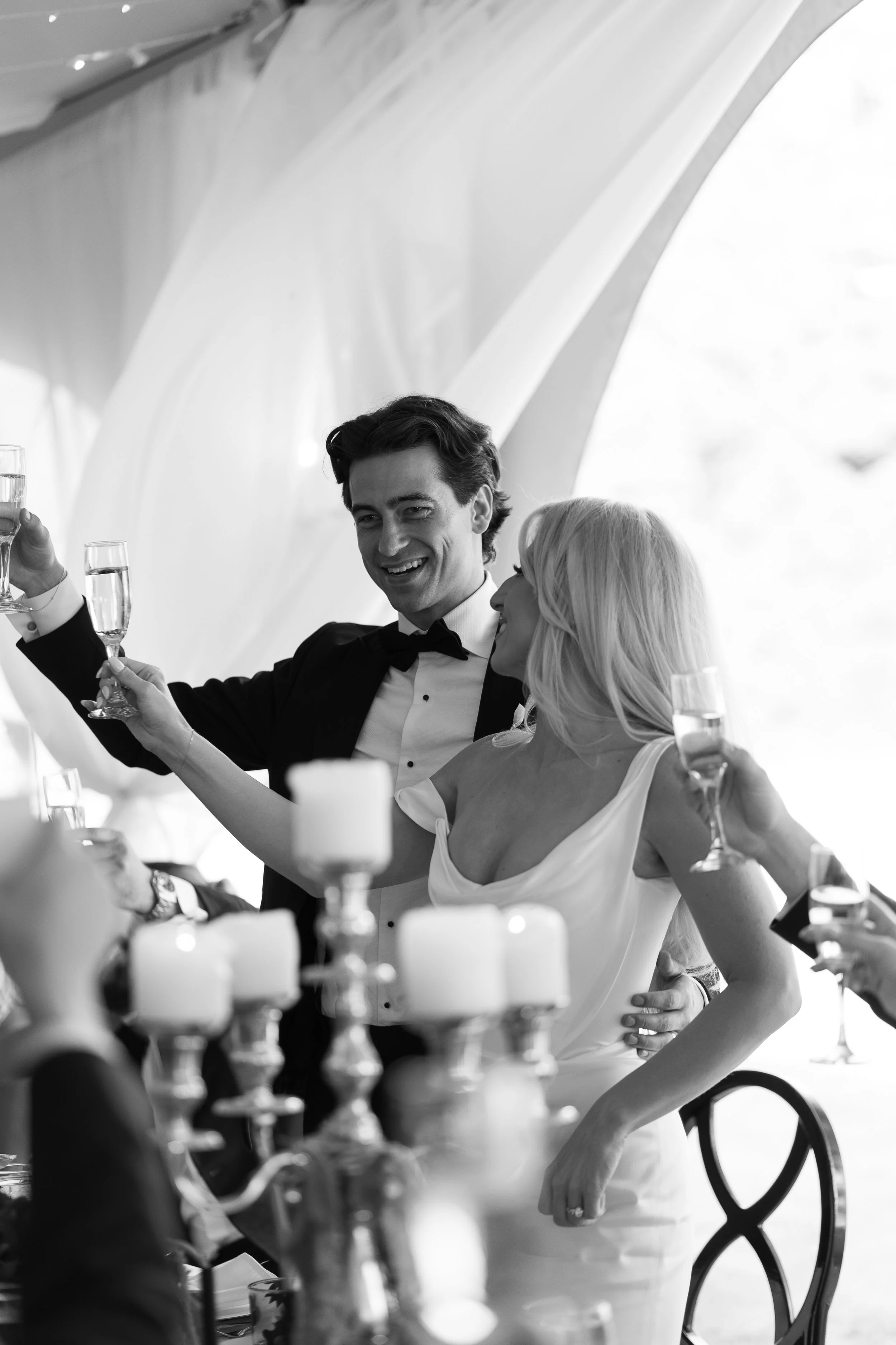 A black and white photo of a man and woman celebrating with champagne at a wedding reception, with candles on the table in the foreground.