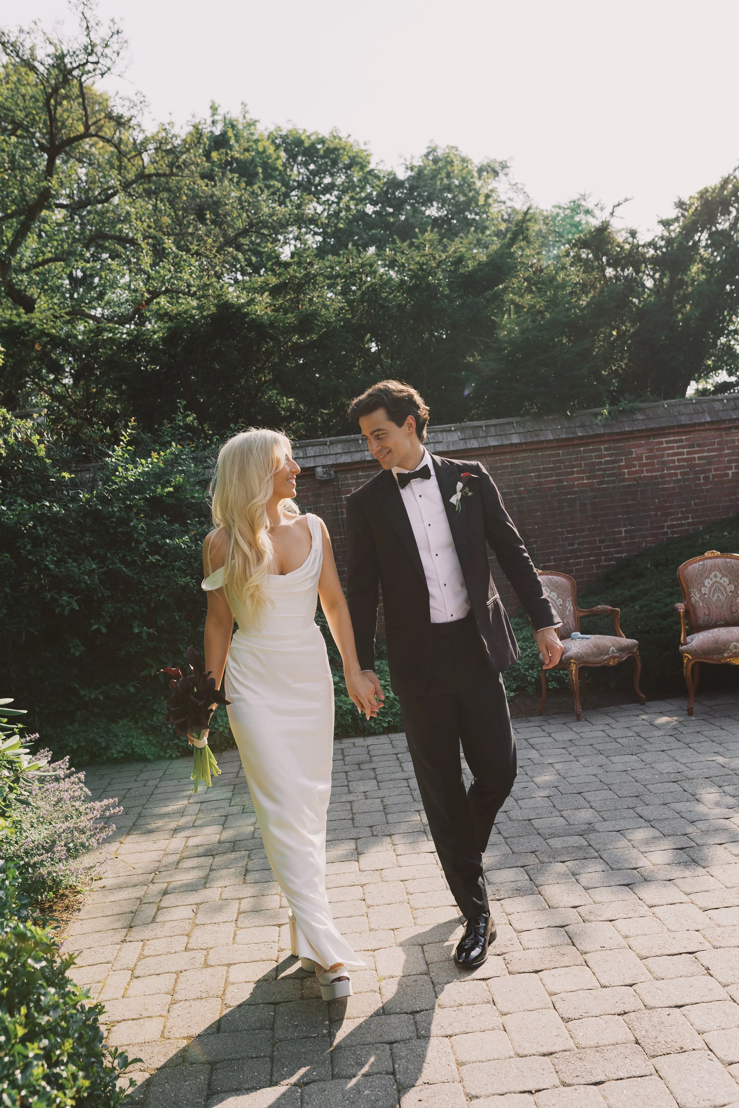A newlywed couple walking outdoors on a patio, holding hands and smiling at each other. The bride has long blonde hair and is wearing a white dress and open-toe heels, holding a bouquet of dark flowers. The groom has dark hair, is dressed in a black 