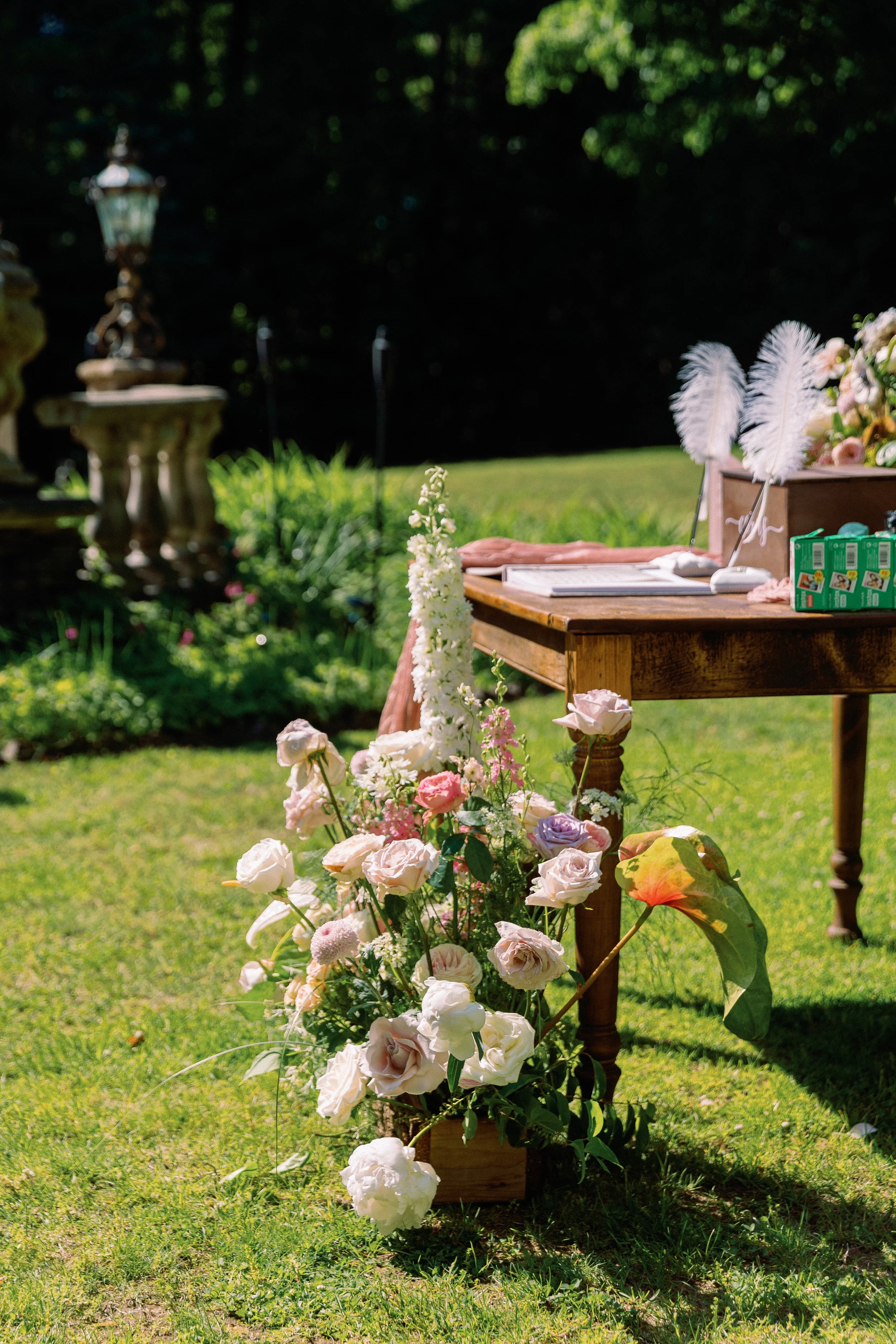 A flower arrangement with roses and other flowers beside a wooden table outdoors, with a garden and stone statues in the background.