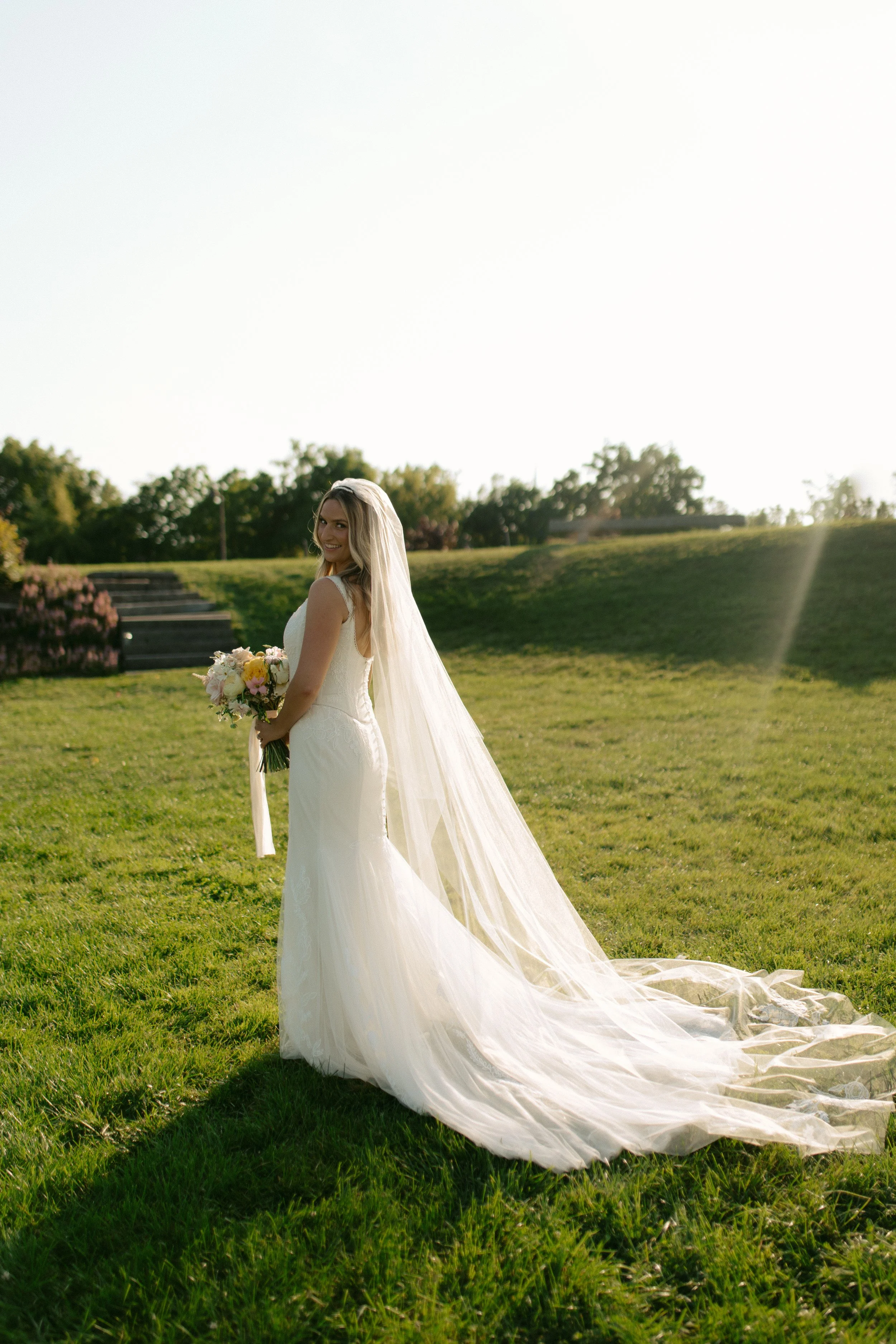Bride in a white wedding gown and long veil holding a bouquet of flowers, standing on grass outdoors with trees and steps in the background, sunlight shining from behind.