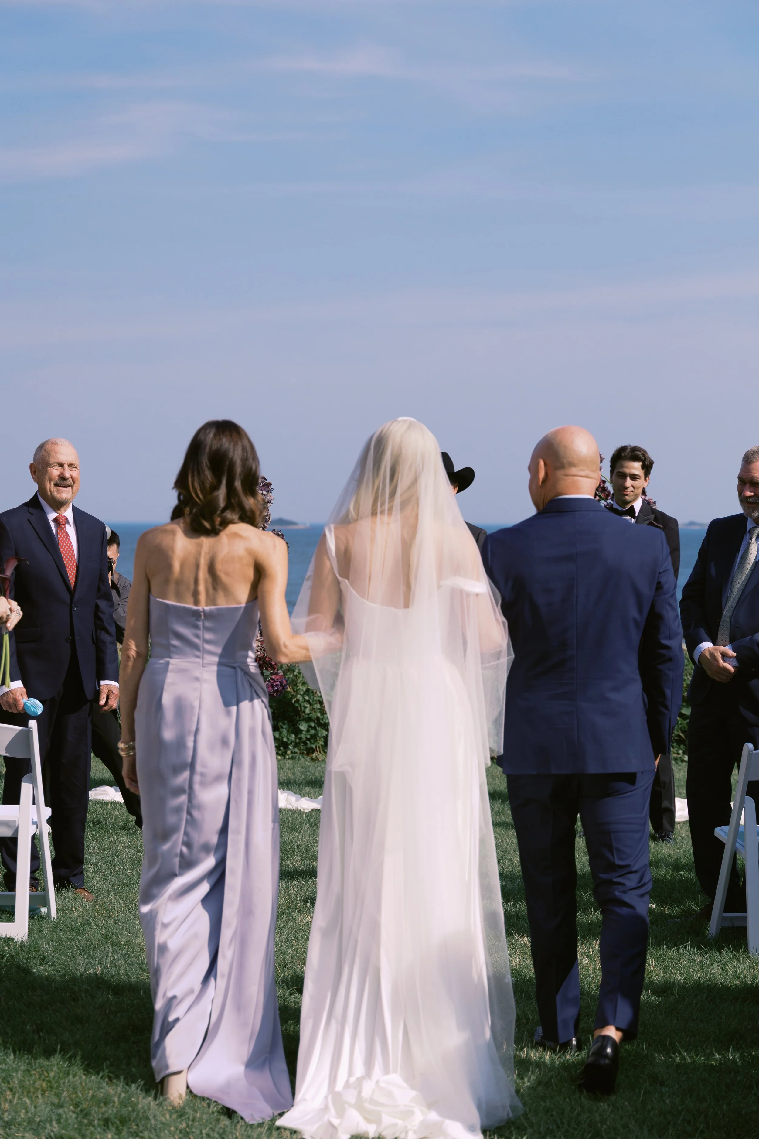 Wedding ceremony outdoors near the ocean with a bride in a white gown and veil, a woman in a lavender dress, and a man in a blue suit, surrounded by seated guests.