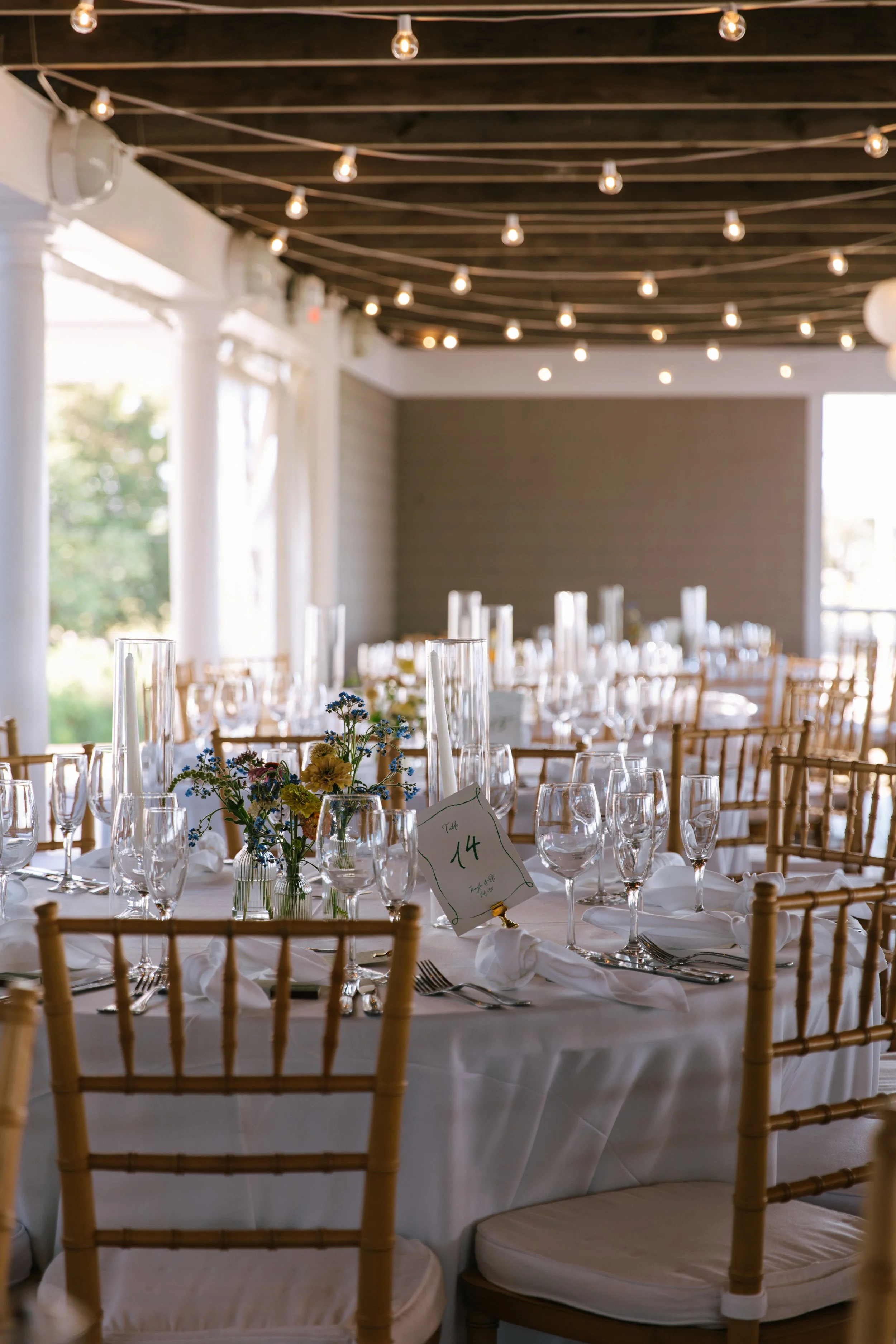 Event space with round tables draped in white tablecloths, decorated with floral centerpieces, glassware, and napkins, set for a formal gathering, with chairs and string lights hanging from a dark wood ceiling.