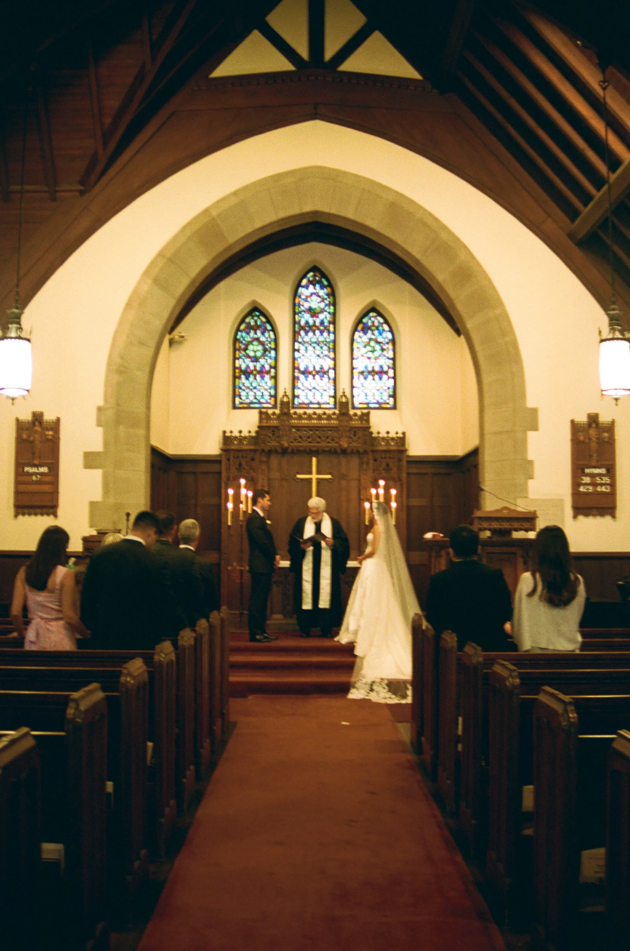 A wedding ceremony taking place in a church with stained glass windows. The bride and groom are standing before a pastor, with guests seated in pews facing them.