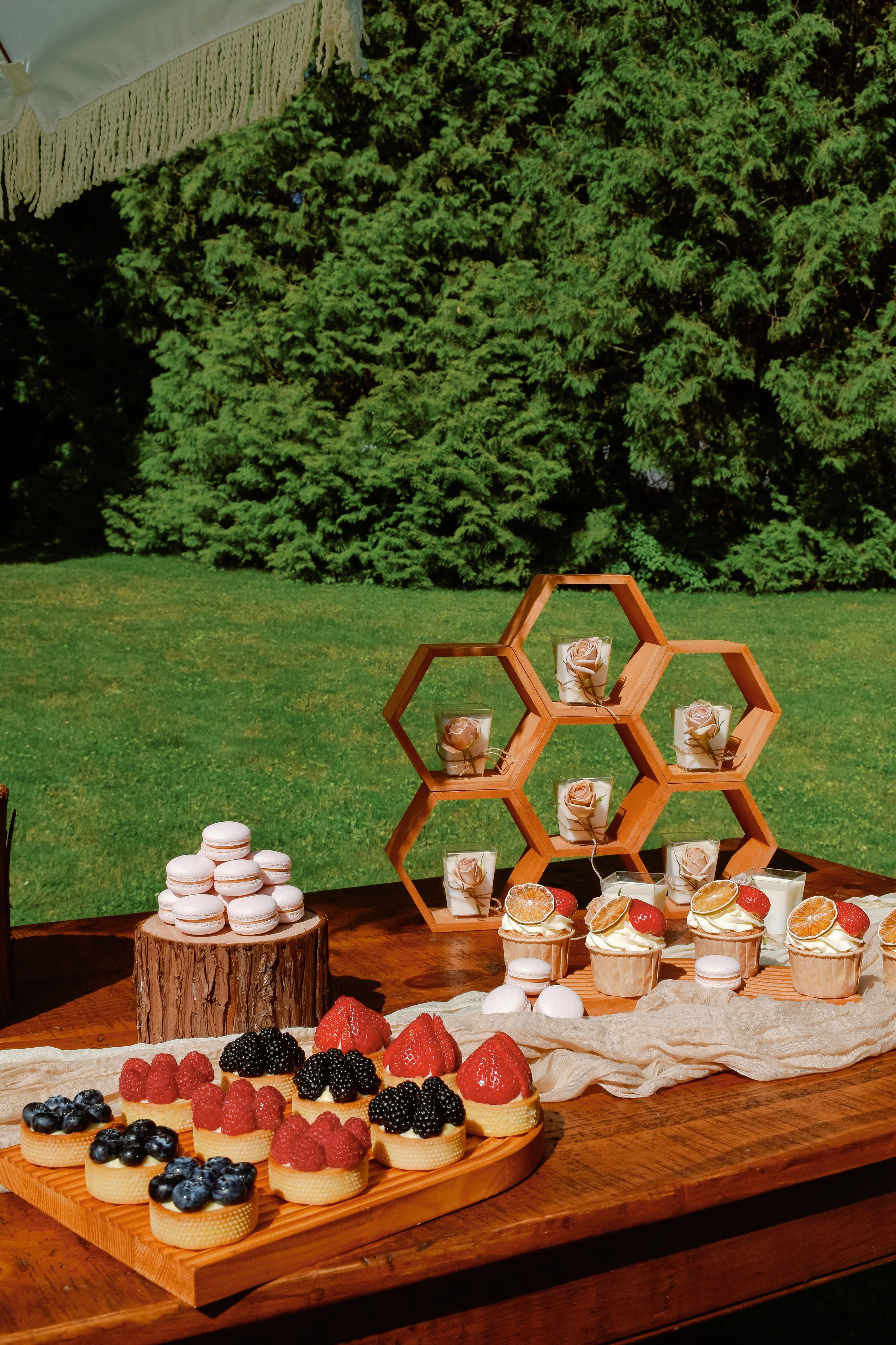 An outdoor dessert table with macarons, berry-topped cupcakes, and fruit tarts, set against a background of green trees and grass.