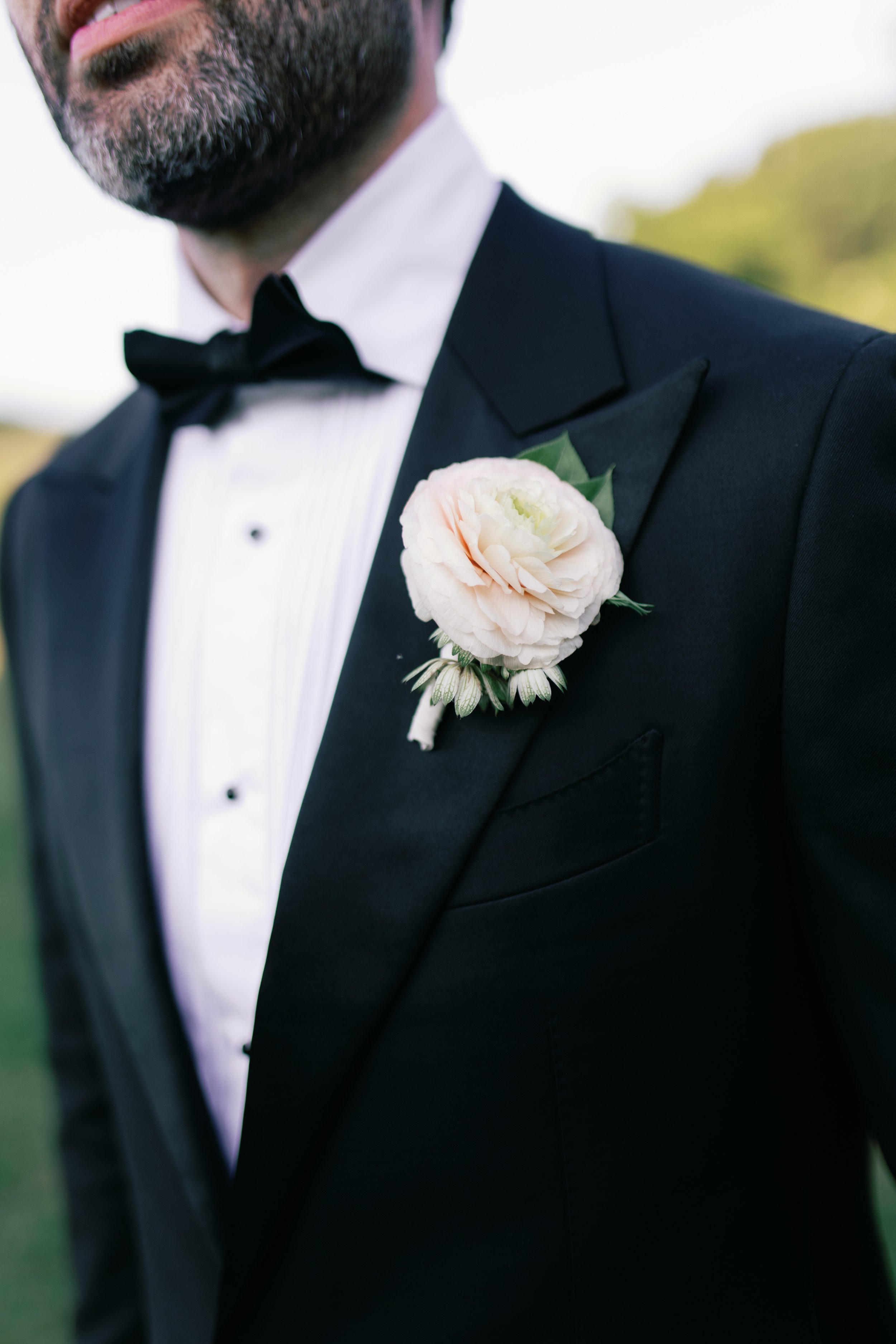 Close-up of a man in a black tuxedo with a white shirt and black bow tie, wearing a white flower boutonniere on his lapel, outdoors with blurred greenery in the background.