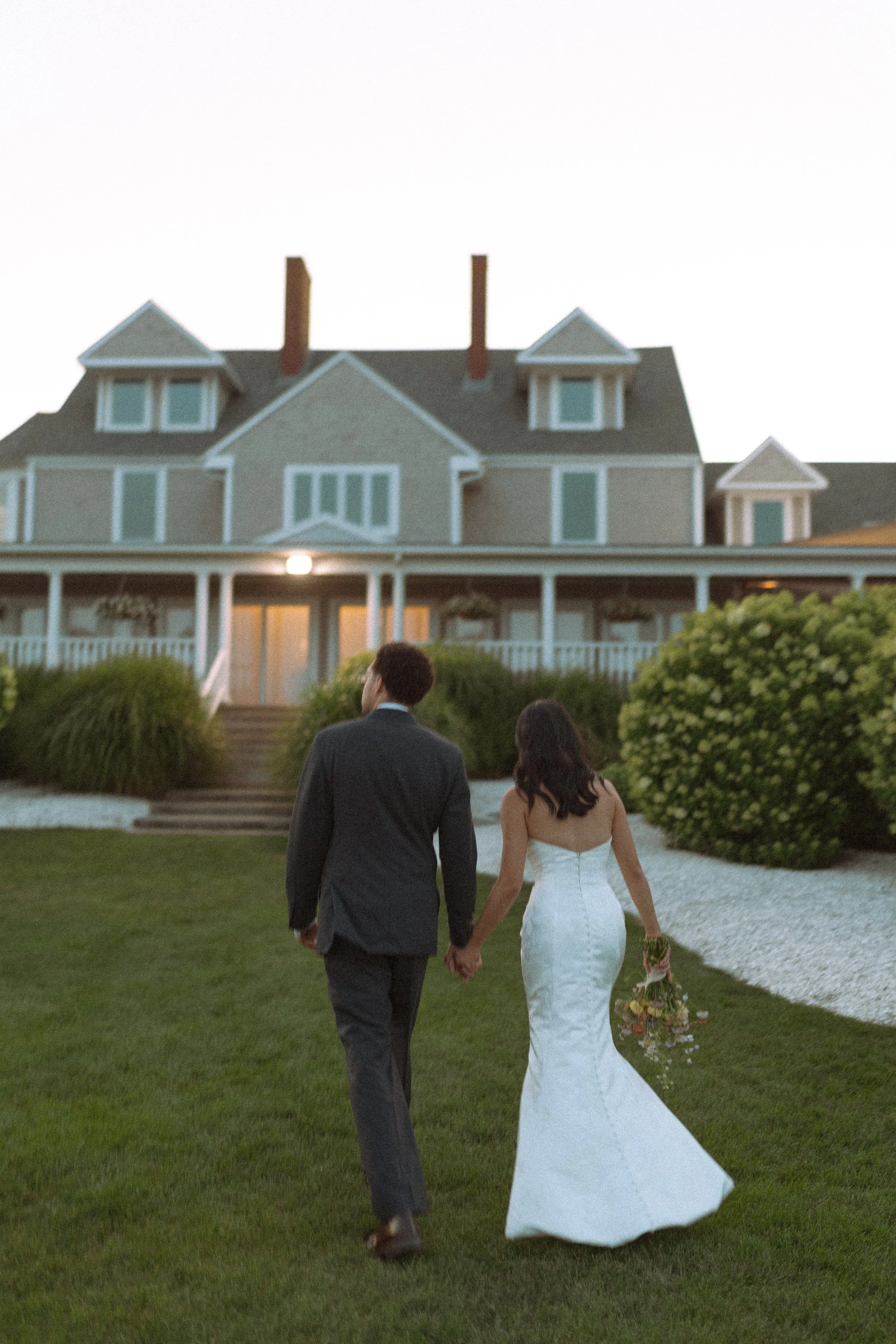 A bride and groom walk hand in hand on a lawn towards a large house at dusk, with the bride holding a bouquet of flowers.