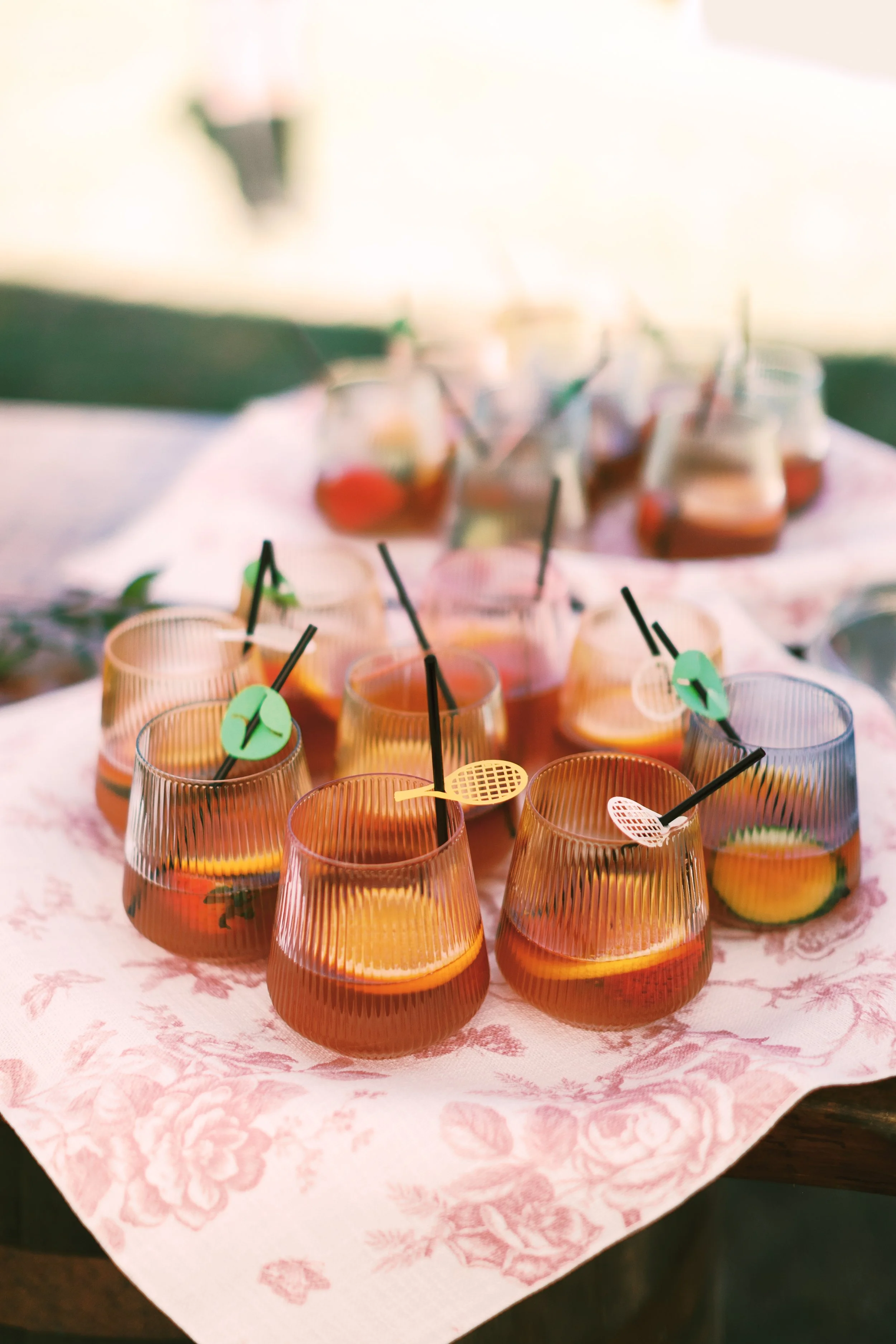 A table with multiple glasses containing a red-colored beverage, each garnished with citrus slices and decorated with colorful stirring sticks.