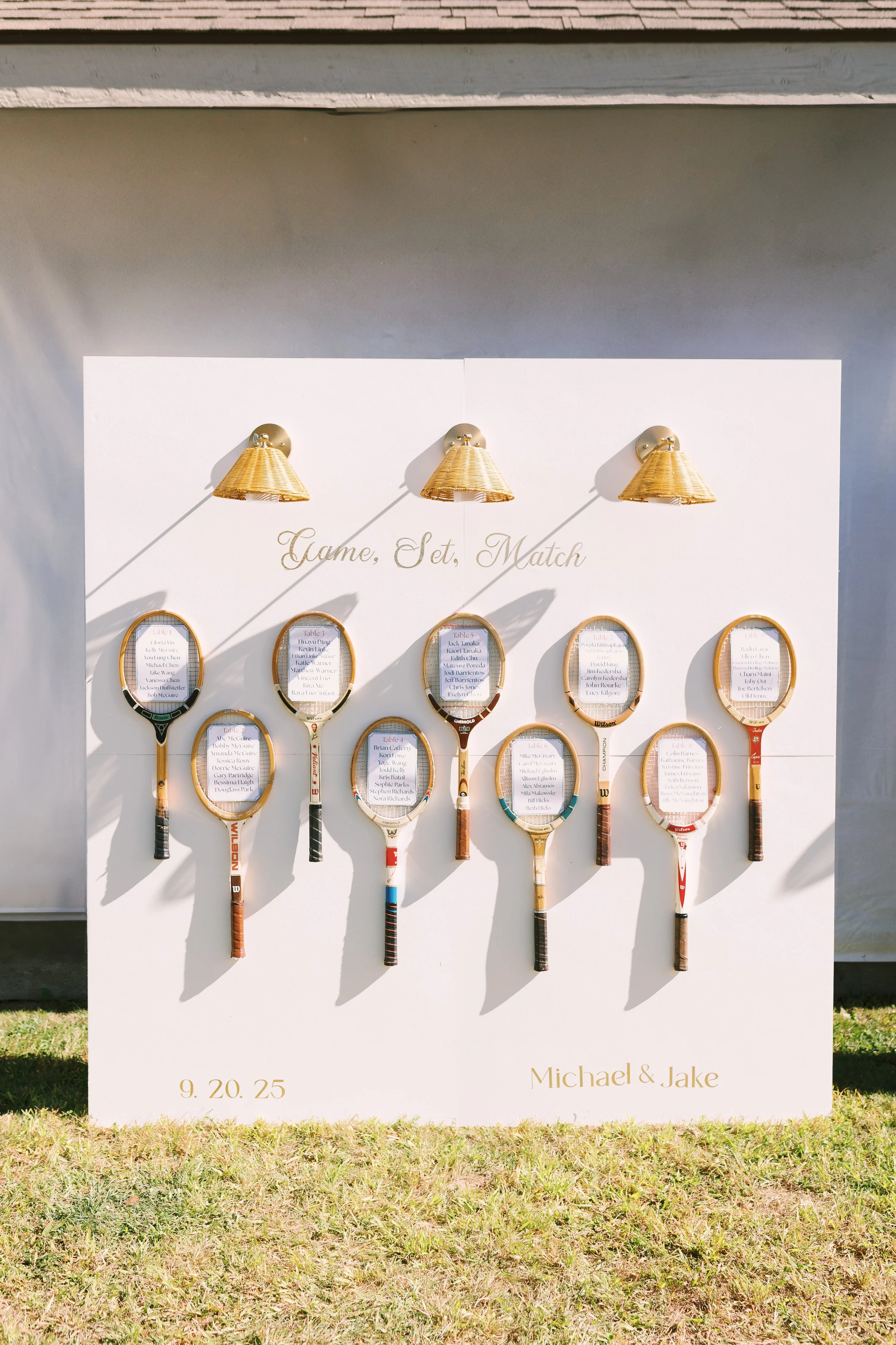 Display board with a tennis-themed seating plan for a wedding. The board has tennis rackets as placeholders for table assignments, with handwritten lists inside each racket. Three gold wall lamps are positioned above the phrase 'Game, Set, Match.' Th