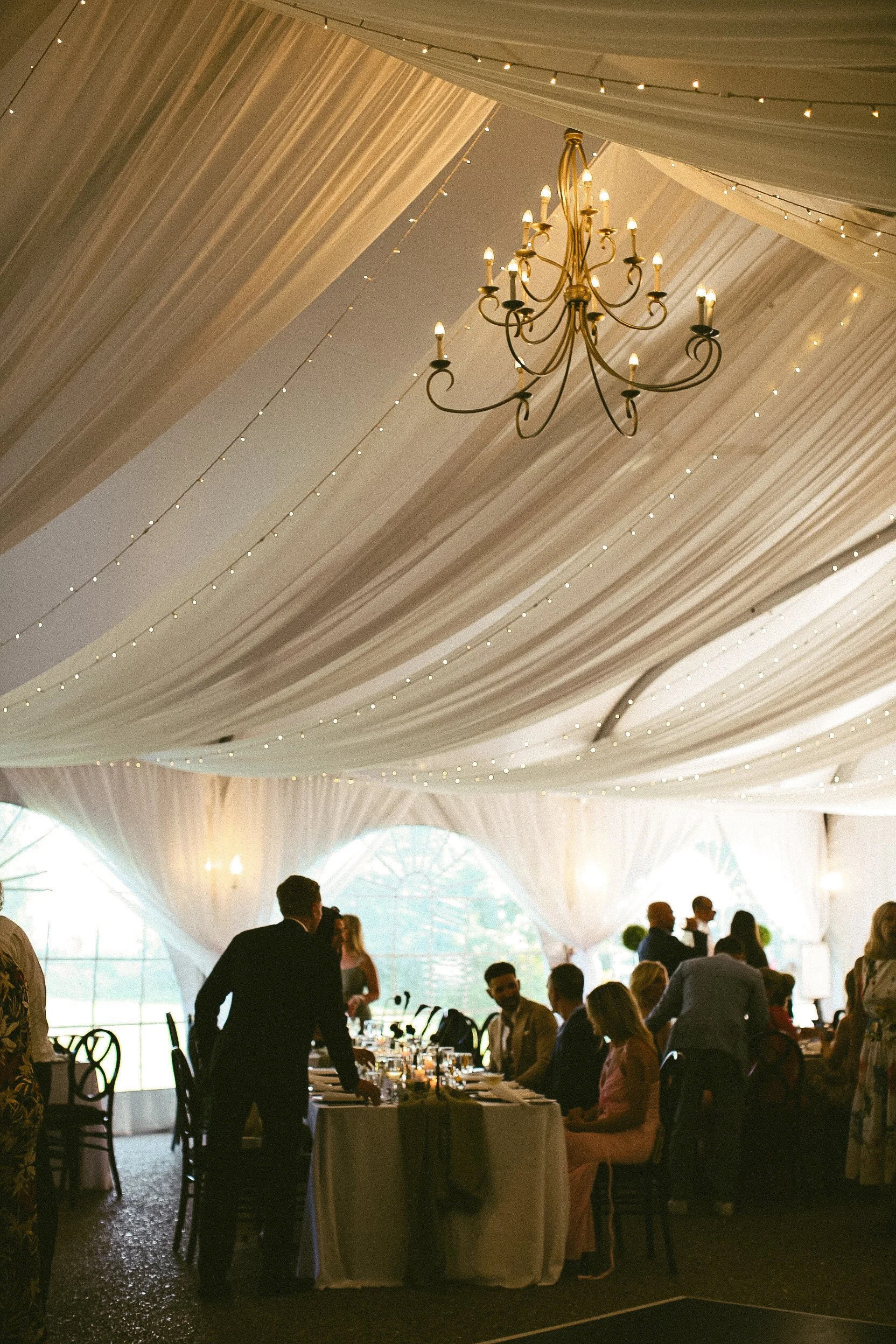 Elegant event tent with draped ceiling, string lights, and a chandelier. People are seated at a long table, with some standing and socializing, during daytime.