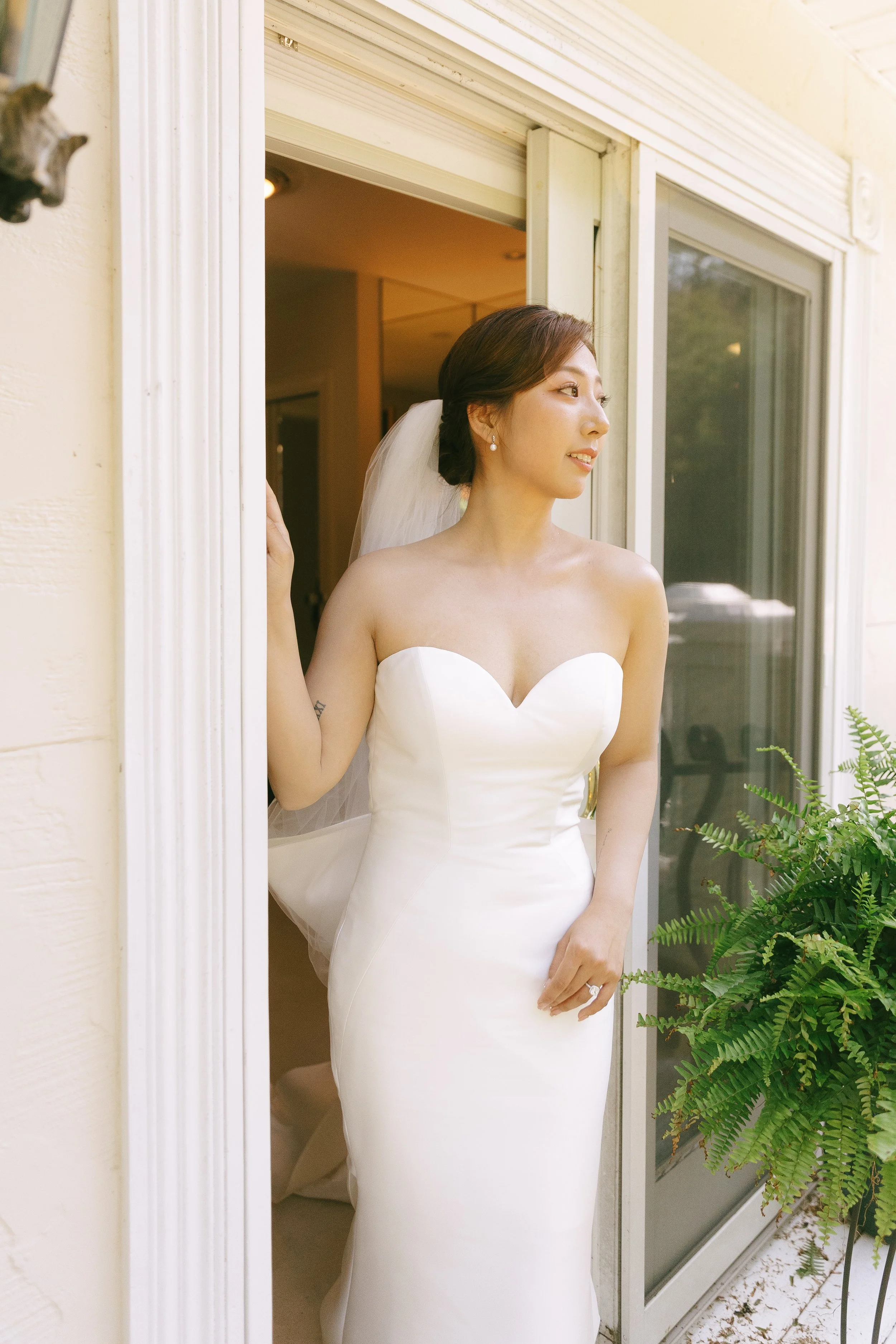 A bride in a strapless white wedding dress, wearing a veil and pearl earrings, stands by a sliding glass door, looking outside with a gentle smile, surrounded by greenery.