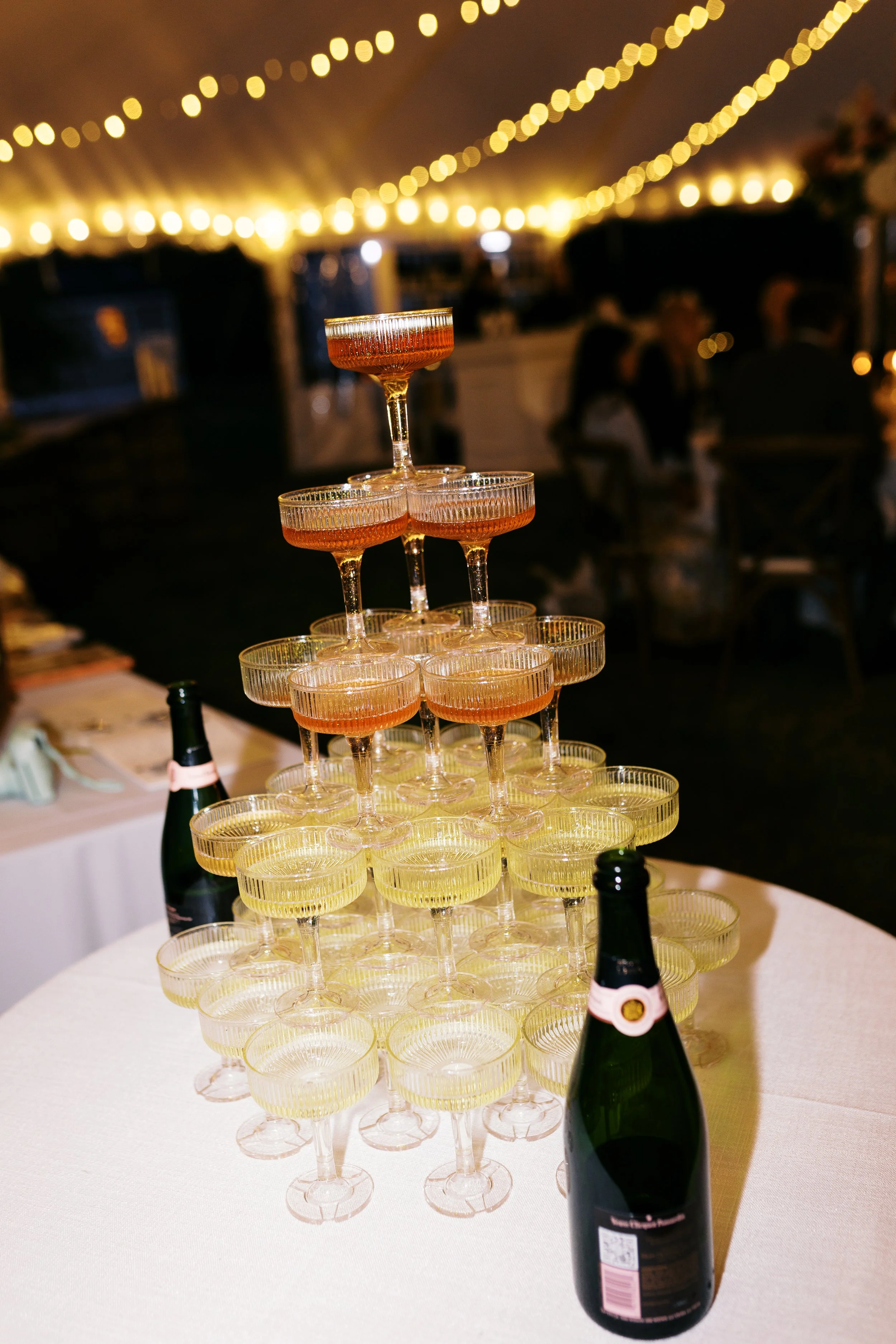 A pyramid of champagne glasses filled with colorful drinks, with two bottles of champagne on a white tablecloth, under string lights at a celebration or event.