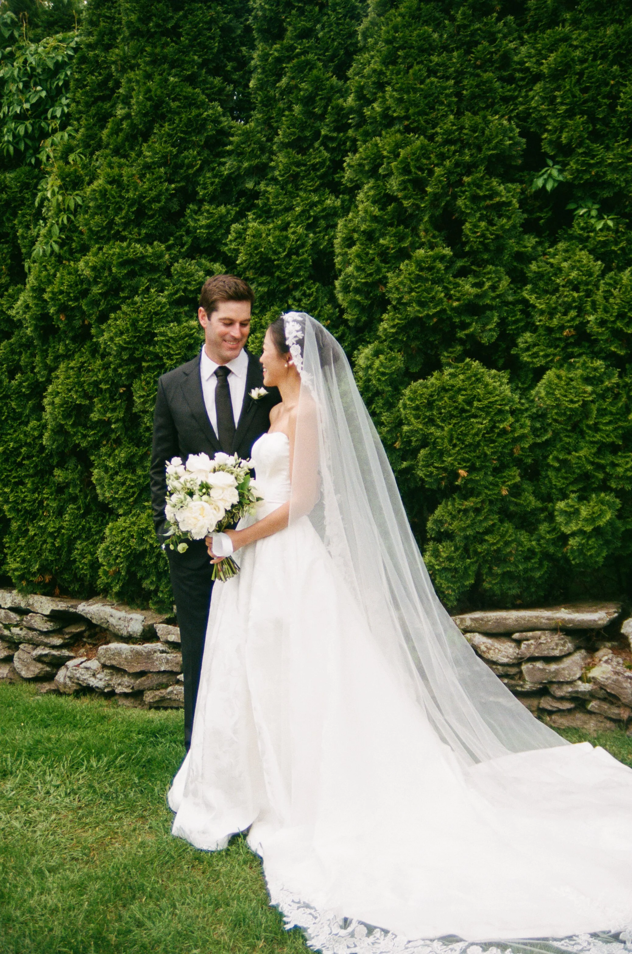 A bride and groom stand outdoors in front of green bushes, smiling at each other. The bride is wearing a white wedding gown with a long veil and holding a bouquet of white flowers. The groom is in a black suit with a white shirt and black tie.