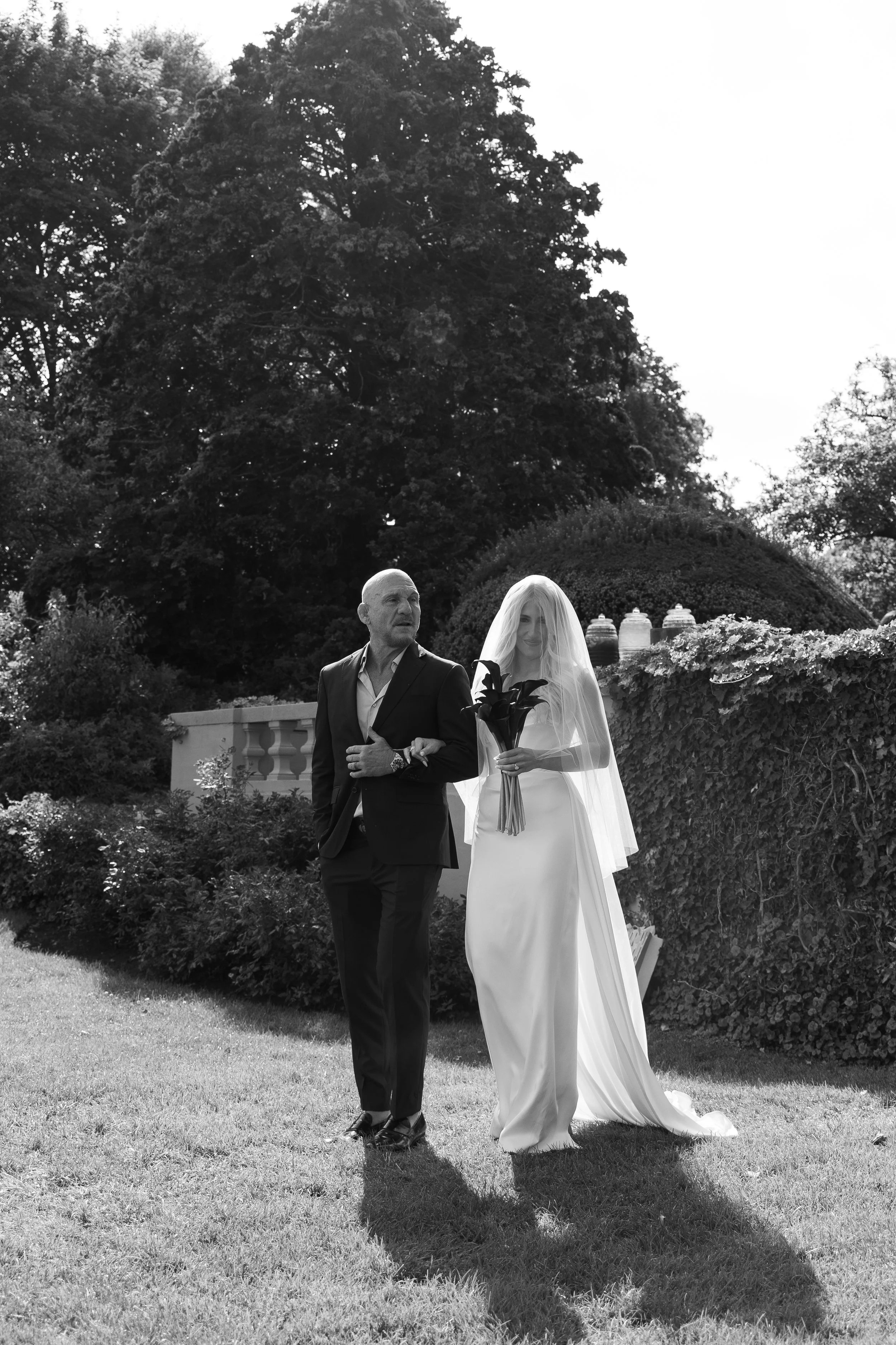A bride in a white wedding dress and veil holding a bouquet walks with an older man in a suit in a garden.