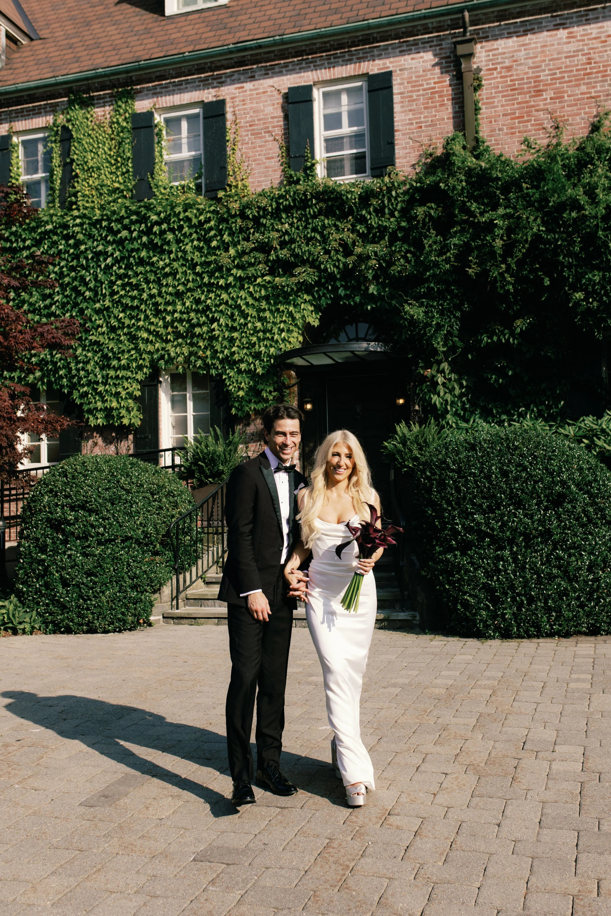 A bride and groom standing outside on a stone pathway, smiling and holding hands. The bride is wearing a white gown and holding a bouquet of dark purple calla lilies, while the groom is in a black tuxedo with a bow tie. Behind them is a brick house w
