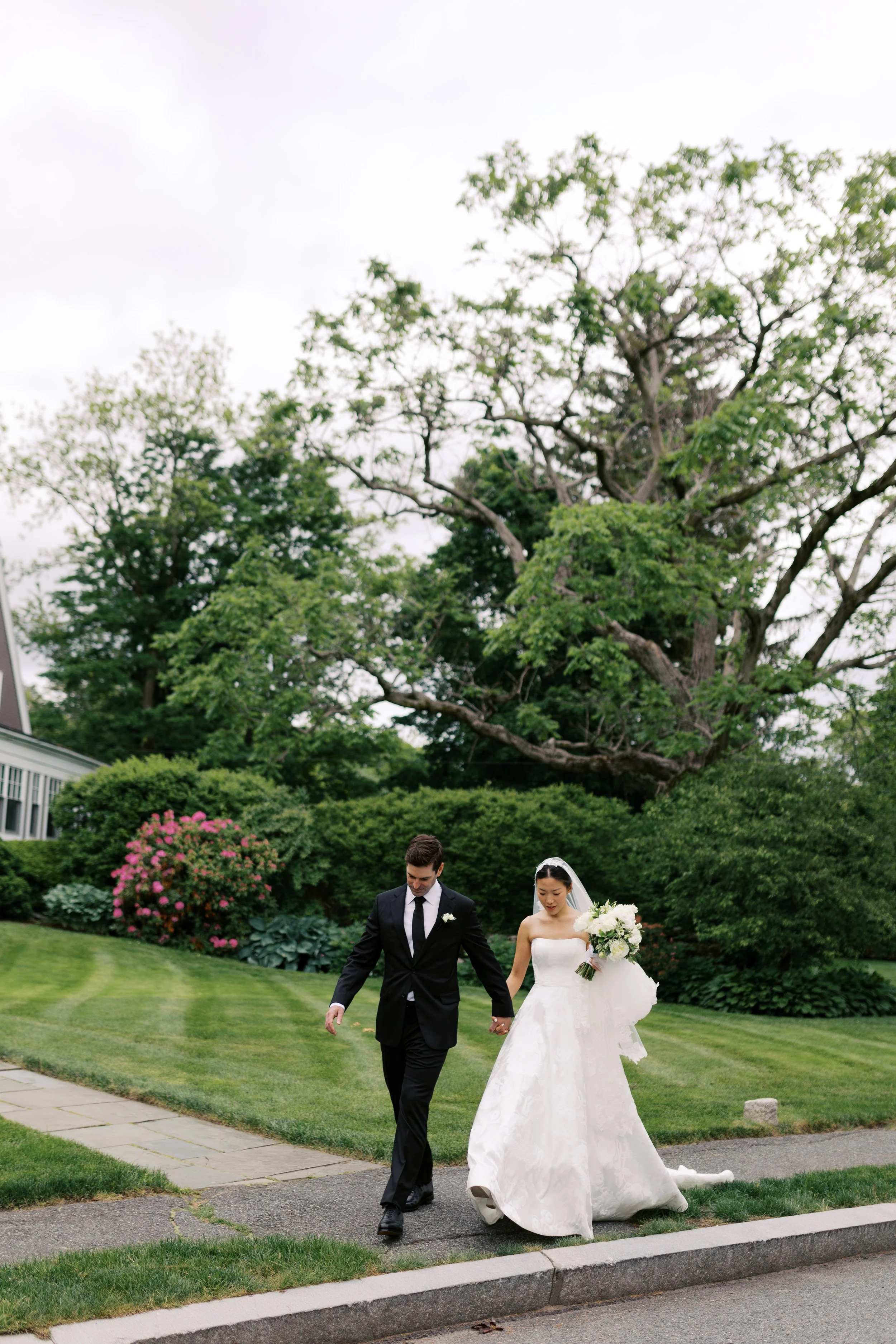 A bride and groom walking hand in hand on a sidewalk in a lush green garden, with the bride holding a bouquet and wearing a white wedding gown, while the groom is dressed in a black suit with a white shirt and black tie.